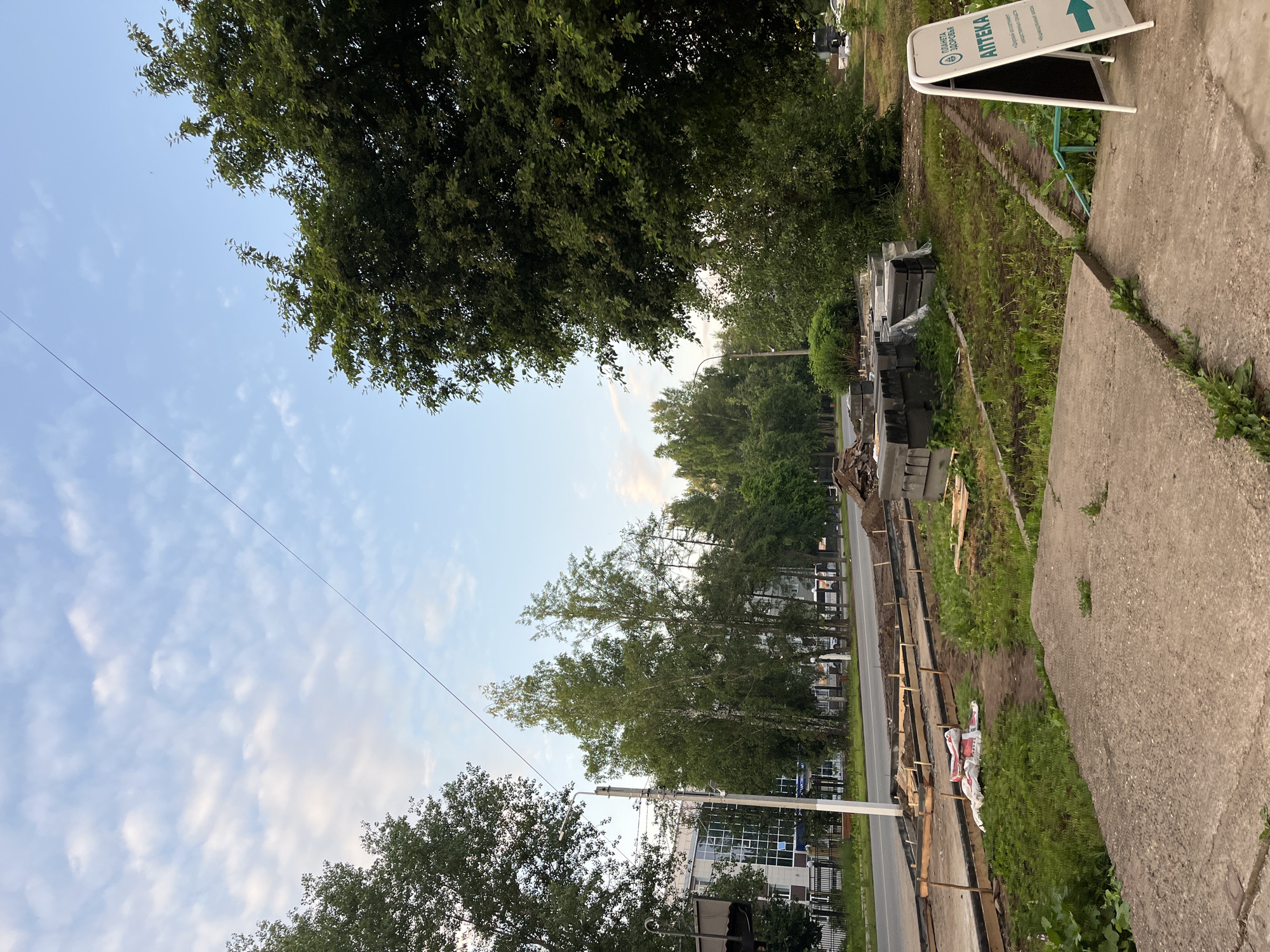 The image depicts a street scene in Chaikovsky, Russia, likely during the daytime with overcast skies. The foreground features a concrete sidewalk and path leading to a road under construction. Several stacks of gray paving stones are visible, along with wooden planks and bags of building materials. A white A-frame sign with Russian text and an arrow pointing right is positioned on the right side of the sidewalk, indicating a pharmacy. Beyond the construction area, a two-lane road is visible with trees lining the side and buildings in the background, including a modern building with a glass facade. The overall scene suggests ongoing urban development or road repairs.
