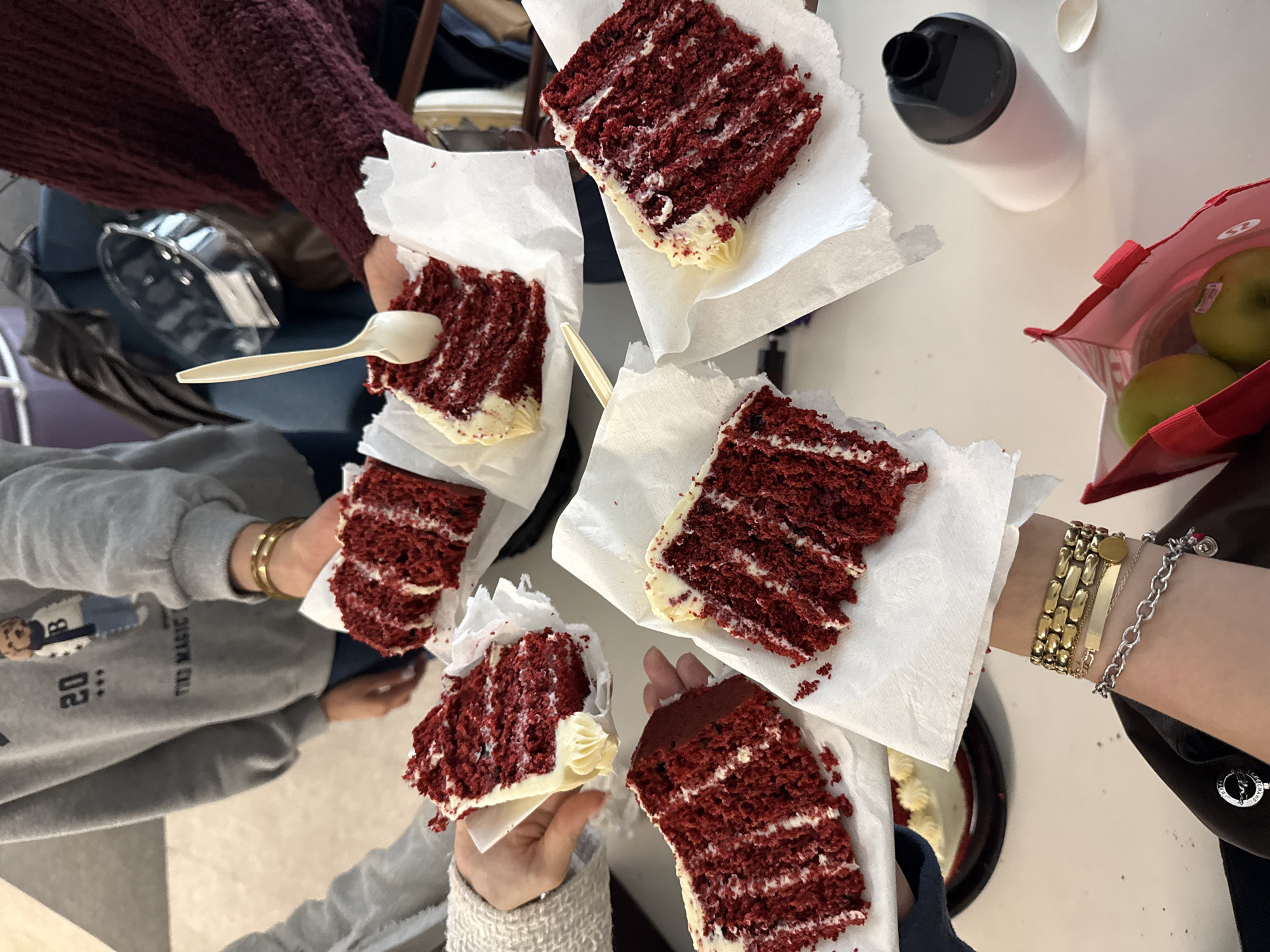 The image shows a group of people holding pieces of red velvet cake on napkins. The camera is positioned overhead, providing a bird's-eye view of the scene. There are five pieces of cake visible, each held by a different person. One person has a plastic spoon in their hand, and another is wearing multiple bracelets. A water bottle and a red bag containing green apples are also visible on the table. The location appears to be indoors, possibly in Mexico City, Mexico, but no specific details confirm this.