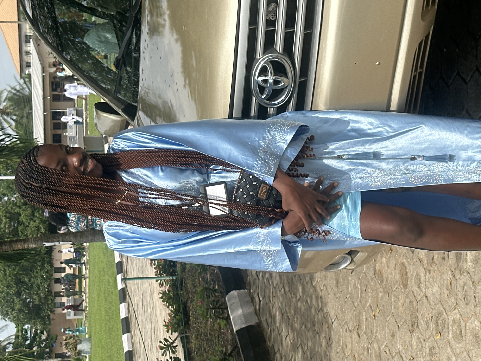 A young Black woman with long, braided hair stands in front of a tan SUV on a paved area. She is wearing a light blue, satin robe with intricate embellishments on the sleeves and hem. Underneath the robe, she is wearing a matching light blue dress. She has a black purse with gold accents hanging from her shoulder, and her hands are clasped in front of her. She is smiling directly at the camera. In the background, there are several people walking on a grassy area, with buildings and trees visible. The scene appears to be outdoors in a relatively bright, sunny environment, possibly on the grounds of an institution or event space in Itoga, Nigeria.