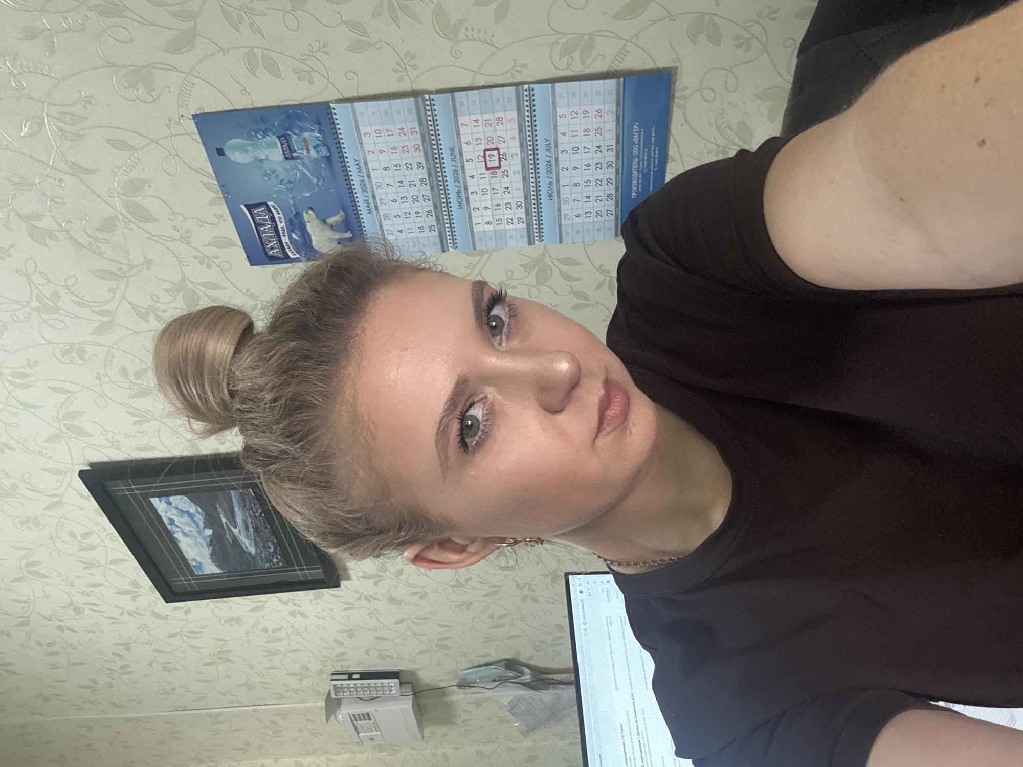 A woman with blonde hair in a bun is shown from the chest up, looking at the camera. She is wearing a dark t-shirt and a gold chain necklace. She has on light makeup with noticeable eyeliner and mascara. The background shows a wall with light green floral wallpaper, a framed picture of a mountain landscape, and a calendar hanging on the wall. The calendar displays the months of May, June, and July of 2026. To the left of the woman, a portion of a computer monitor and a white electronic device are visible.