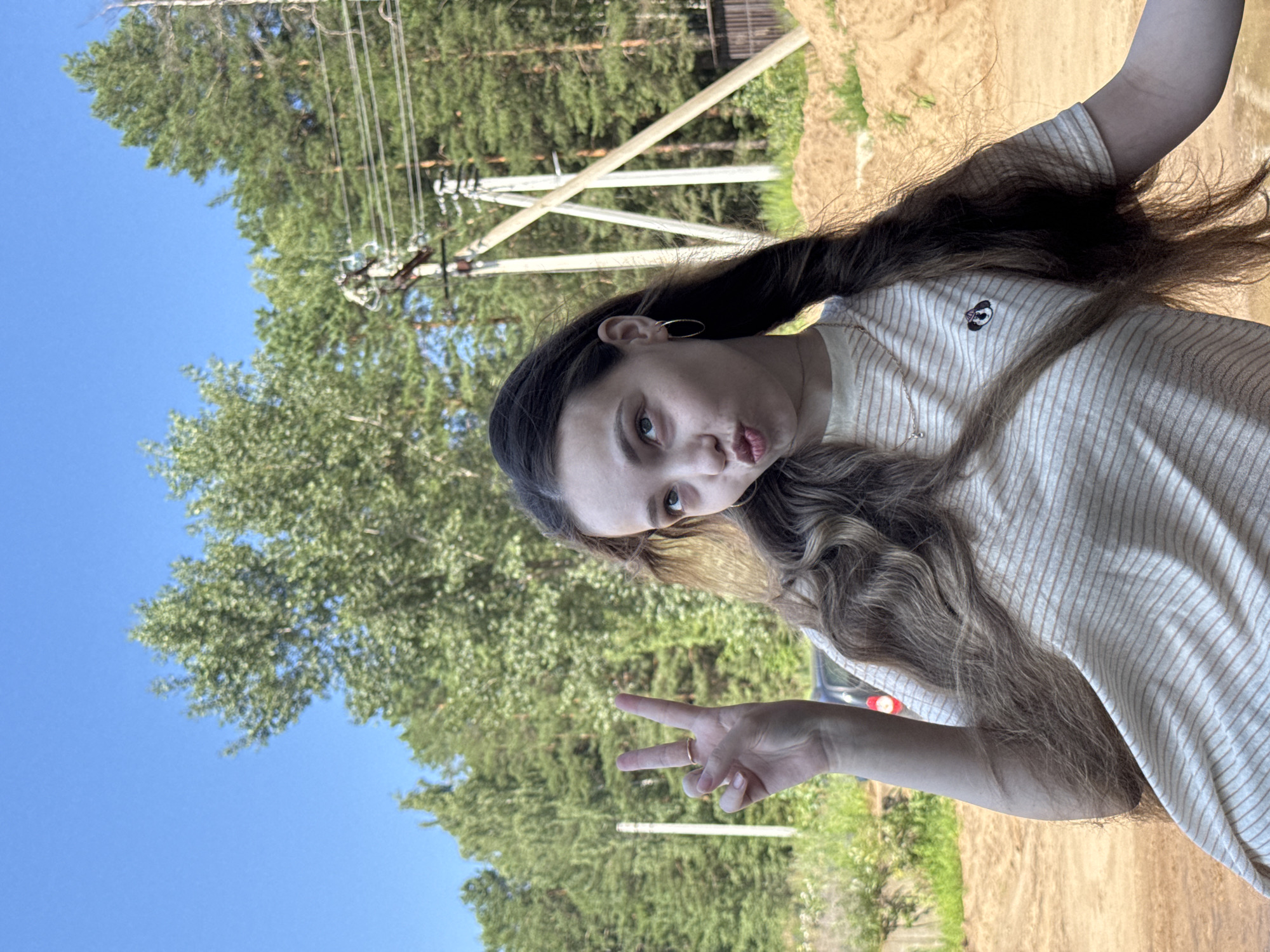 The image shows a young woman with long, wavy brown hair, wearing a striped t-shirt. She is outdoors, with a clear blue sky and a dense forest in the background. There are power lines and utility poles visible. The ground appears to be dirt and sand. The woman is making a peace sign with her left hand and puckering her lips. The scene suggests a rural or suburban setting in Noviy, Russia, likely during daylight hours given the bright conditions.