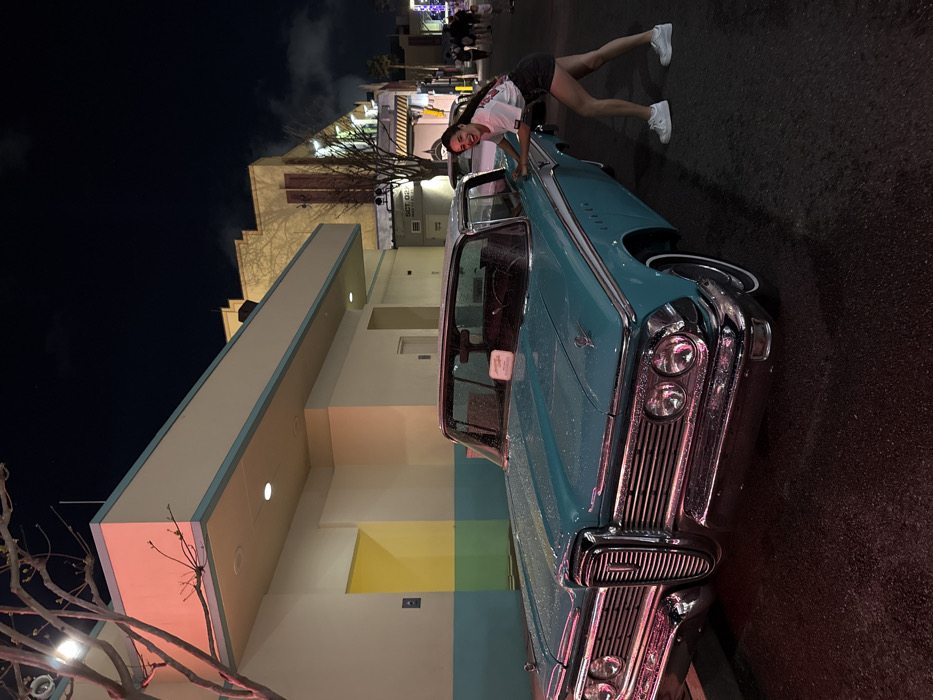 The image captures a nighttime scene in Orlando, Florida, specifically within a themed entertainment area consistent with Universal Studios. The primary focus is a vintage two-tone automobile, identifiable as a 1959 Edsel in turquoise with a white roof, positioned on a wet asphalt street. Its chrome grille, stacked headlights, and bumper are prominent, reflecting red and white artificial lighting. Water droplets are visible on the vehicle's surface.
A young adult female is depicted to the right of the car, playfully leaning into its passenger side rear fender with both hands, adopting a pushing stance with her right leg bent. She has dark hair, is smiling towards the camera, and is dressed in a white graphic t-shirt, dark shorts, and white sneakers.
The background consists of buildings exhibiting a mid-20th-century architectural style. To the immediate left, a cream-colored building with a light blue stripe and a recessed yellow alcove is visible, partially obscured by a dormant tree. Further along the street to the right, additional structures feature stepped, geometric facades. The sky is dark, indicative of nighttime, and artificial lighting illuminates the scene, creating reflections on the wet pavement. In the distant background on the right, several indistinct figures suggest the presence of other individuals.