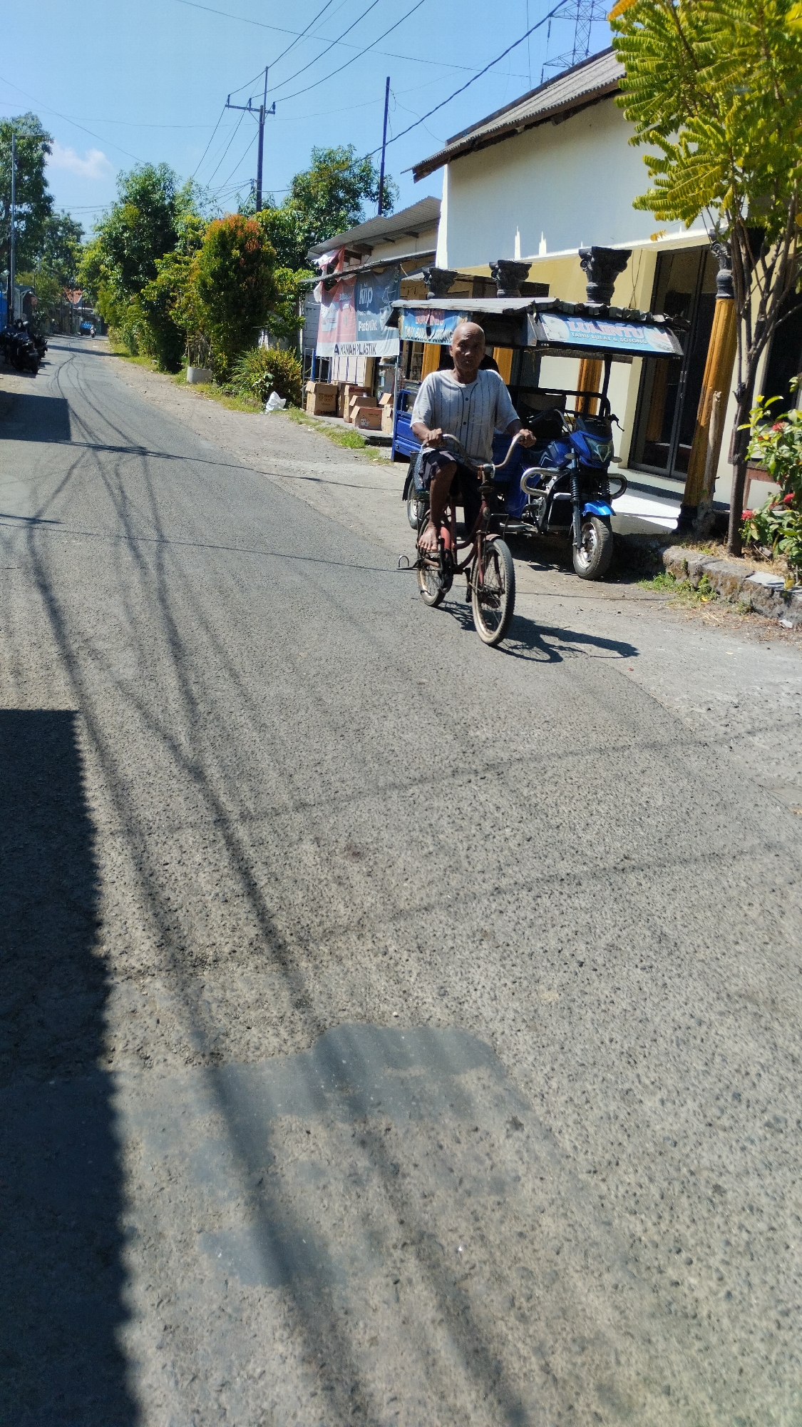 The image captures a street scene in Kramat Jegu, Indonesia, on a bright, sunny day. In the foreground, an older man with a bald head and wearing a striped short-sleeved shirt is riding a red bicycle. The bicycle appears to be a child's or a small adult bicycle, with white tires and chrome handlebars. The man is looking forward, and his feet are positioned on the pedals. To the right of the bicycle, a blue motorcycle is parked. The street is paved with asphalt and shows signs of wear. Numerous tree shadows are cast across the road from overhead power lines and unseen trees. In the background, there are buildings, including a white building with a tiled roof and a smaller structure with a corrugated metal roof. Several trees with green foliage are visible along the side of the road, and some orange and red flowers are growing near the white building. To the left of the street, parked motorcycles are visible. Signs are posted on the buildings in the background, with some text visible but not clearly legible. The sky is a clear blue.