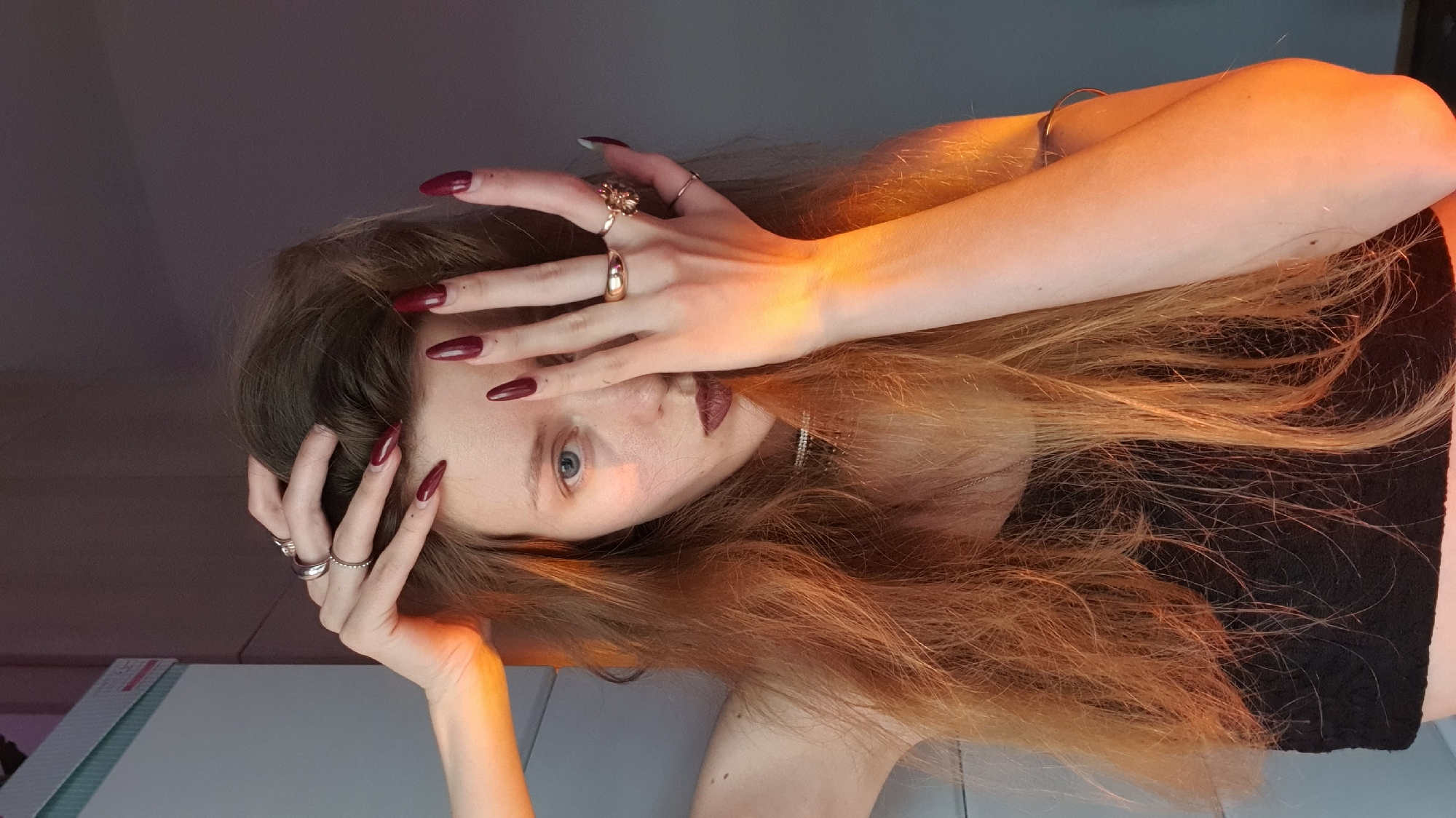 A woman is seen in a close-up shot, lying down with her head tilted slightly to the right. Her hair is long, reddish-brown, and tousled, partially obscuring her face. She has fair skin, light blue eyes, and dark lipstick. Her hands are positioned on her face and hair, adorned with multiple rings. She is wearing a black top. The lighting is uneven, with a warm, orange hue on one side and cooler tones on the other. The background is a gradient of dark to light gray. There is no indication of location, including the city of Bar, Montenegro, in the image.