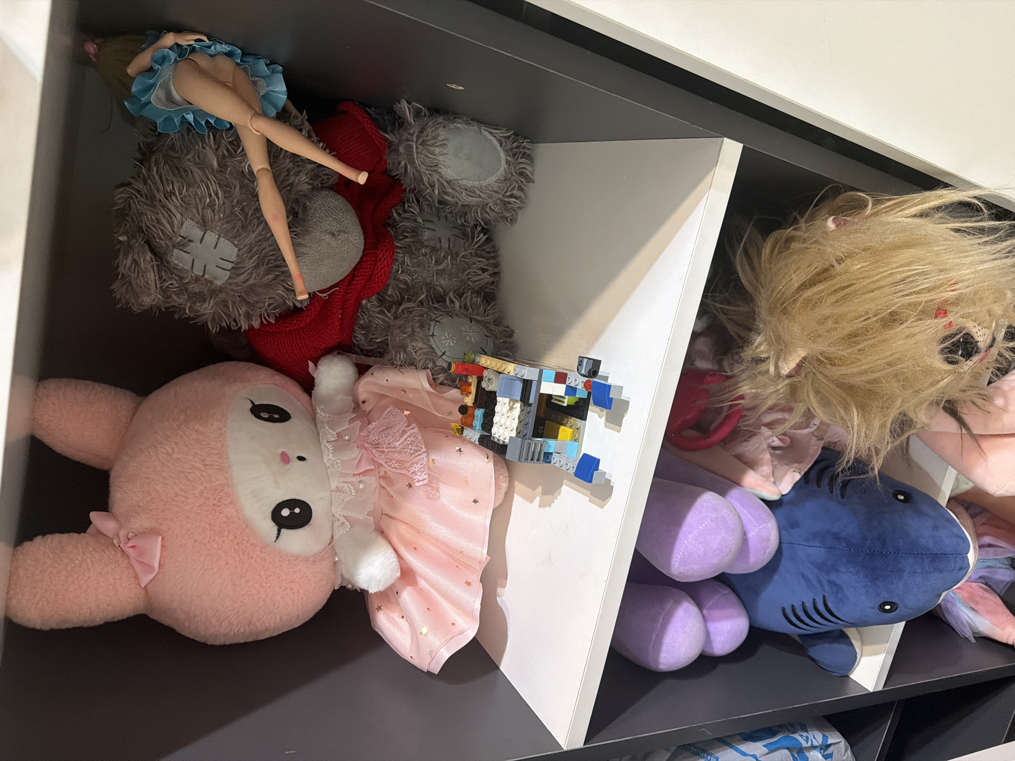 The image shows a shelf filled with stuffed animals and a small LEGO structure. On the top shelf, a large pink bunny plush toy in a sparkly pink dress sits next to a grey teddy bear wearing a red sweater. A doll with a blue skirt is positioned behind the teddy bear. A LEGO structure is on the shelf in front of the stuffed animals. The shelf below contains a blue shark plush toy, a purple stuffed animal, and other plush toys that are partially visible. The environment appears to be an indoor storage space, possibly a closet or a shelf in a room. The image does not provide any information to determine its location in the city of Shymkent, Kazakhstan.