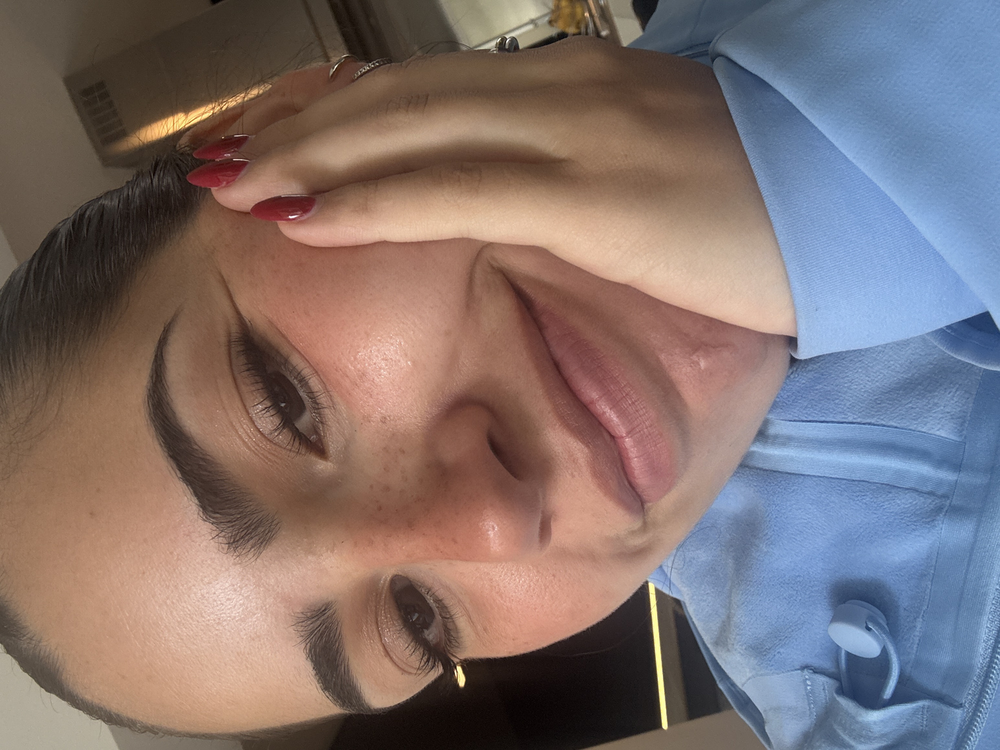 The image is a close-up selfie of a woman, taken from a slightly high angle. She has dark hair pulled back, prominent eyebrows, and is wearing light makeup, including mascara, eyeshadow, and lip gloss. Her skin has a natural tone with some freckles visible on her nose and cheeks. Her right hand is placed on the left side of her face, with bright red nail polish on her fingernails. She is wearing a light blue hooded jacket with a drawstring toggle. The background is slightly out of focus, showing a hint of a modern interior with a radiator and some lighting. There is no indication of the location being in San Bernardino Tlaxcalancingo, Mexico.