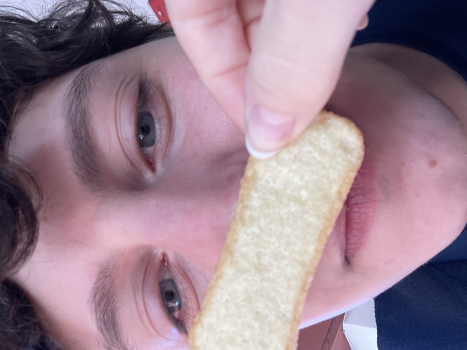 A close-up, diagonal portrait features the upper face of a person, likely young, with dark, curly hair framing their face. Their skin has a light complexion, and their prominent eyes are a light blue or grey color, with visible dark pupils. The eyebrows are dark and well-defined.

A hand, with short, light-colored fingernails, holds a long, rectangular, light-golden biscuit or cookie, possibly a ladyfinger, directly in front of the person's mouth. The biscuit appears to be plain, with a slightly textured surface. The person's lips are slightly parted, hinting at readiness to eat or having just taken a bite.

The shot is tightly framed, focusing on the interaction between the person and the biscuit. The background is largely out of focus, with hints of a dark blue garment on the right side of the frame and a lighter, possibly white, surface on the left, suggesting an indoor setting. The image is oriented diagonally, giving it a dynamic, candid feel, possibly a selfie. The person's expression is neutral, primarily focused on the biscuit. No text is visible in the image. This photo was taken in Dosso (Ciappanico), Italy.
