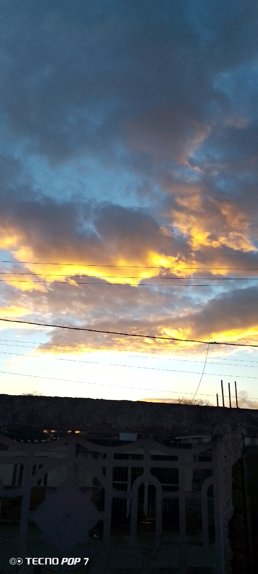 The image is a low-angle shot of a sunset sky with clouds colored in orange and grey, seen above a cityscape. A network of power lines cuts across the sky. Below the horizon line, there is a dark silhouette that appears to be a building or a structure with a few vertical posts extending upward. In the foreground, there's a fence made of concrete or stone, partially obscured by shadow. The bottom left corner of the image displays "TECNO POP 7." The image was taken in the city of Altagracia, Venezuela.