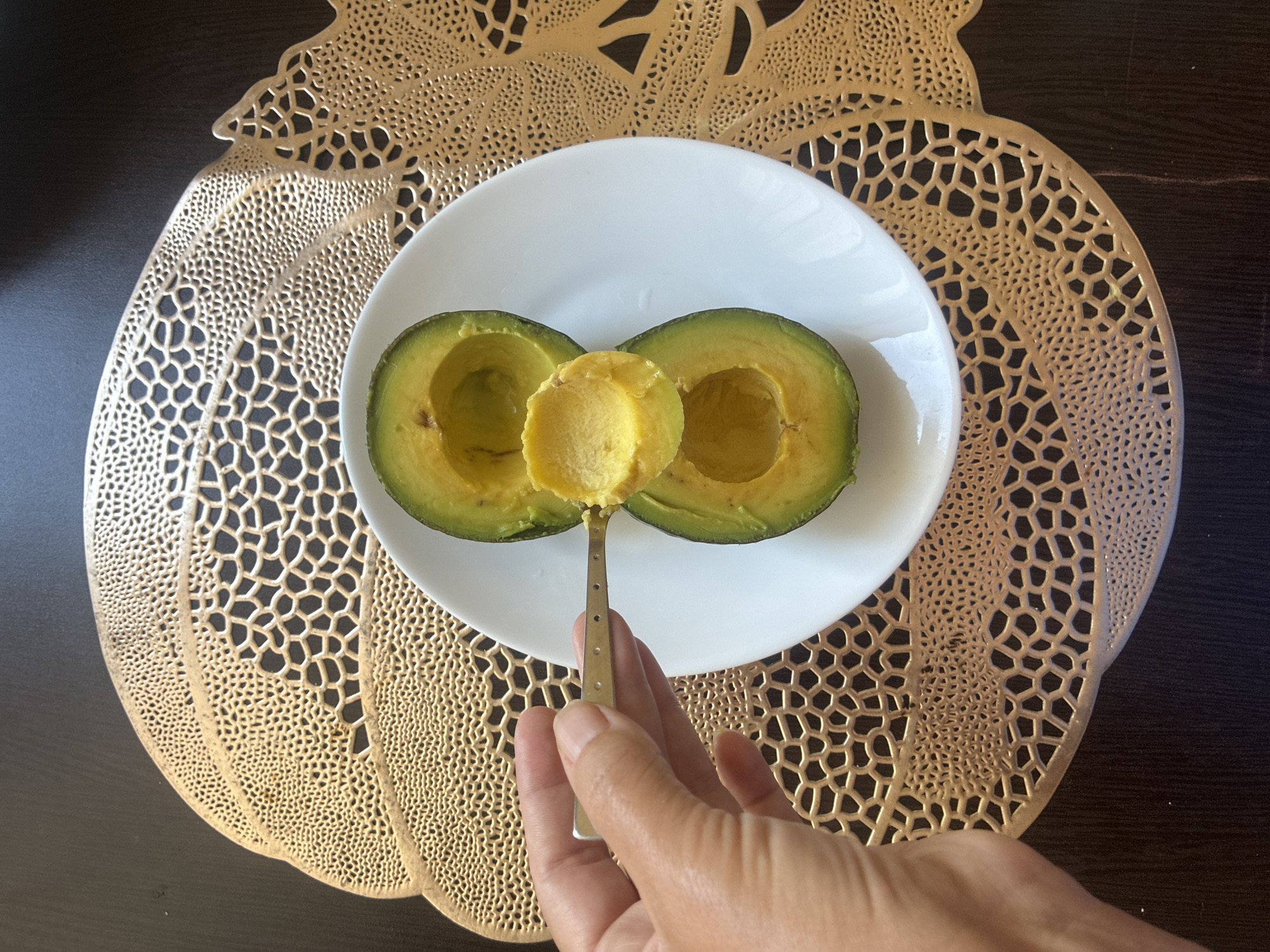 The image shows a person's hand holding a small spoon, scooping a piece of avocado from a halved avocado that rests on a white plate. The plate is positioned on a decorative, gold-colored placemat with an intricate, honeycomb-like pattern. The placemat is placed on a dark, textured surface, likely a table. The background is dark and out of focus. The avocado is green on the outside and yellow-orange on the inside, with one half showing more flesh scooped out than the other. The lighting suggests an indoor setting, possibly a kitchen or dining area, with natural light casting shadows. There is no indication of the city of Sochi, Russia, in the image.