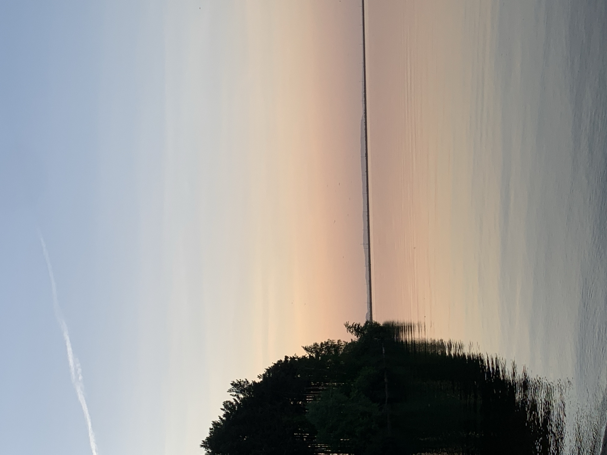 The image captures a serene waterscape under a pastel sky. The colors transition from a pale blue on the left to peach and soft orange hues towards the center and right, suggesting either sunrise or sunset.
The horizon line features a narrow dark strip, possibly indicating land or distant objects. Reflections are visible on the water's surface, mirroring the colors of the sky and the dark shapes on the horizon. A forested shoreline occupies the bottom of the frame, with the reflection of the trees clearly mirrored on the water. A contrail is visible on the left side of the image.
Given the request for the city of Chaikovsky, Russia, the scene likely depicts the Kama River or one of its reservoirs, which are prominent features of the landscape in that area. Without further context, specific details about people, actions, or interactions are not discernible in the scene.