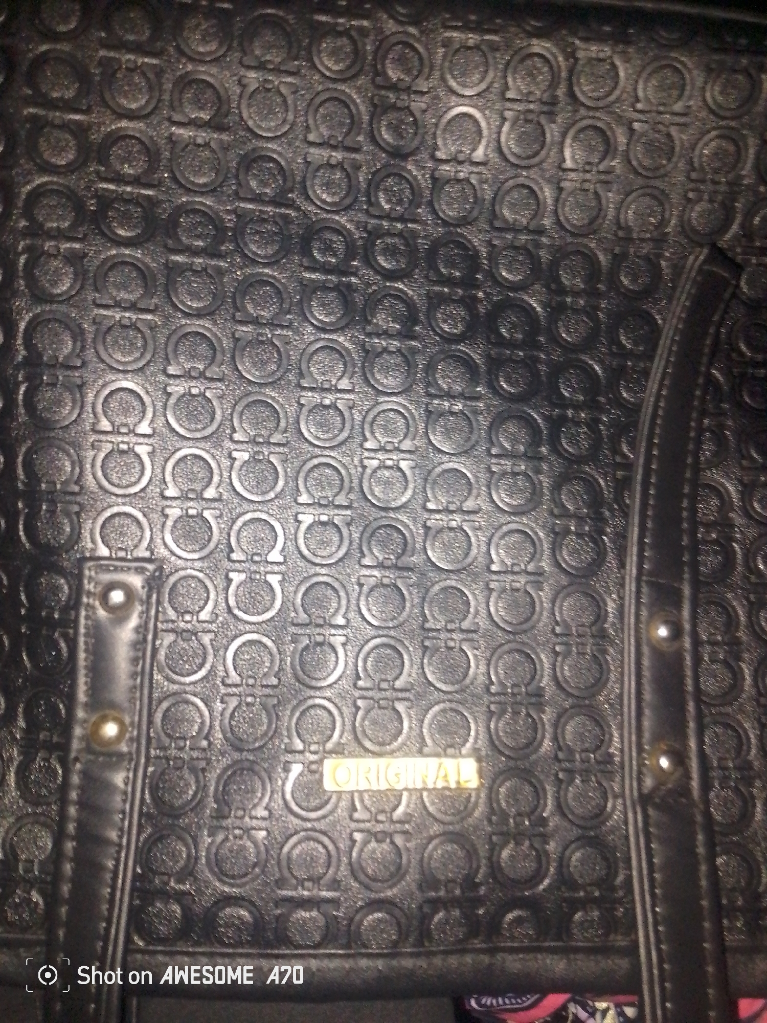 The image is a close-up, low-angle shot of a black leather handbag with an embossed pattern of interlocking "G" shapes. The handbag has a textured surface and appears to be a tote bag style with visible stitching and hardware details, including rivets and a metallic "ORIGINAL" logo. The lighting is dim, creating shadows that highlight the embossed pattern. The bottom left corner of the image shows a watermark indicating "Shot on AWESOME A70." The scene is indoors with no discernible urban environment visible.