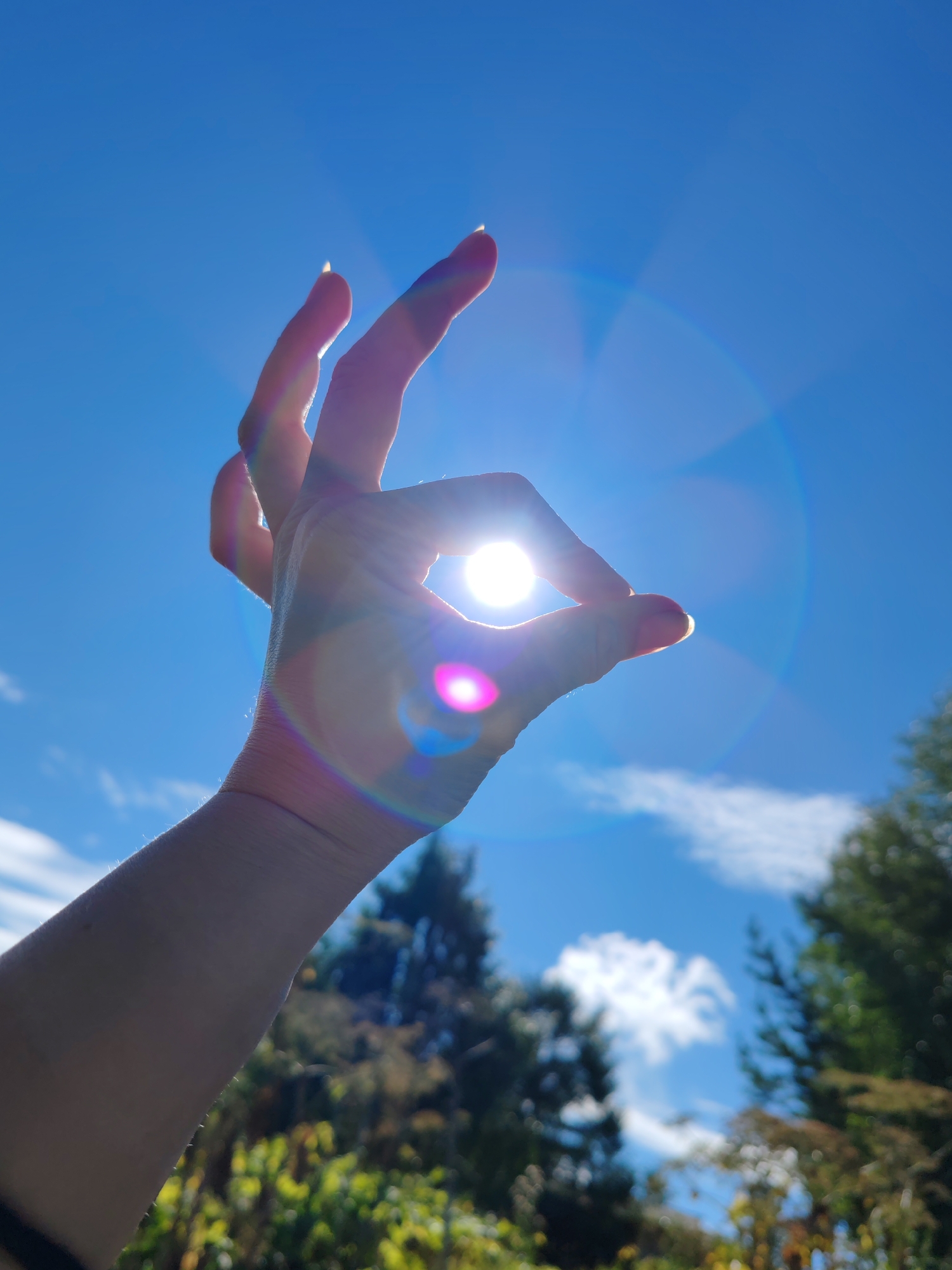 A hand is held up against a bright blue sky, with the sun shining through the circle formed by the thumb and index finger. The sun appears as a bright, white orb, and there are lens flares and light streaks visible around it, creating a rainbow effect. The hand is in focus, with its palm facing the viewer, and the fingers are slightly spread. In the background, the sky is clear with a few scattered white clouds. Below the sky, there is a blurred landscape of trees and foliage, suggesting an outdoor setting, possibly in a park or natural area. The specific location in the city of Чухлэм, Russia cannot be determined from the image alone.