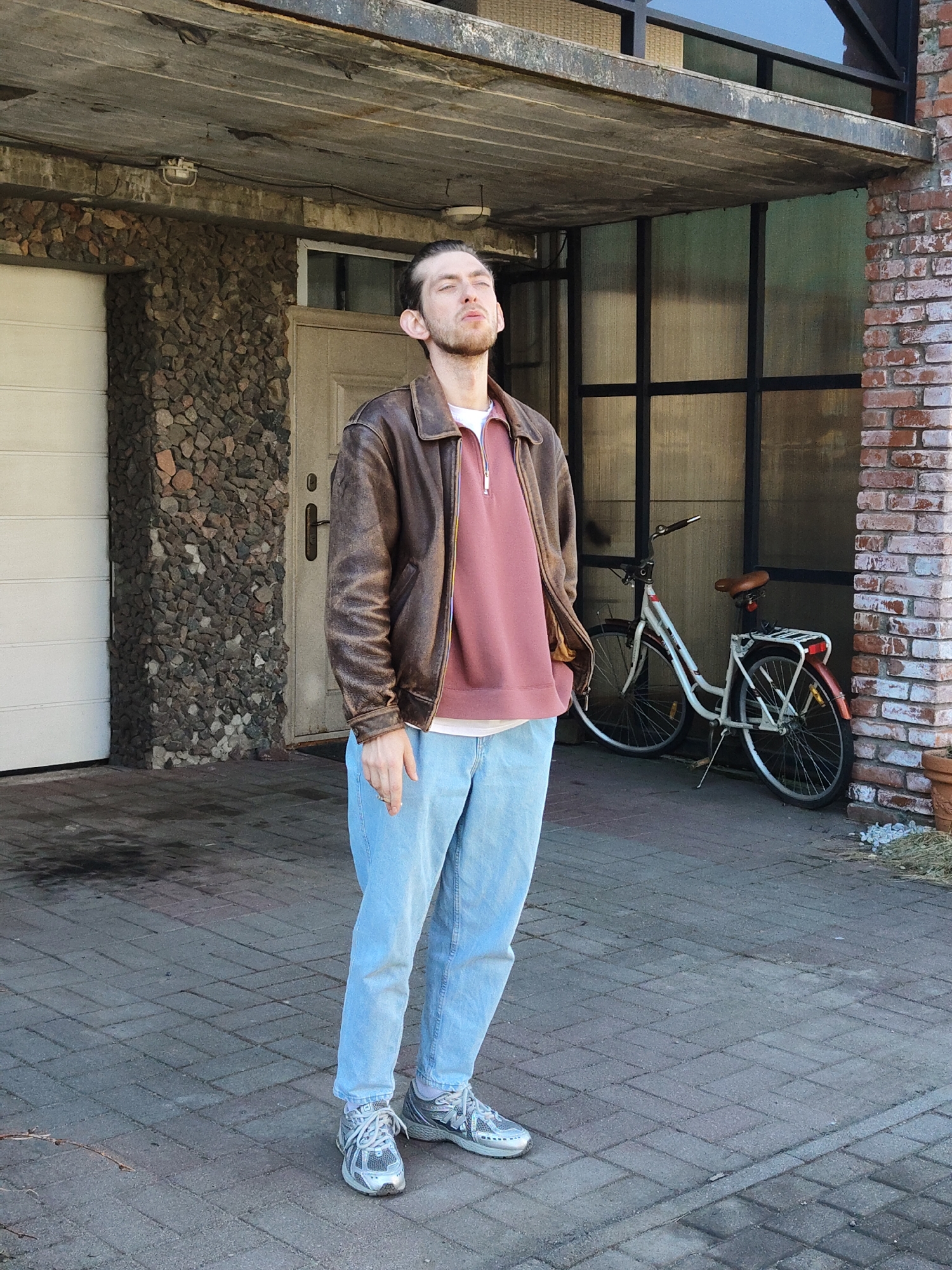 A full shot captures a Caucasian male standing in front of a building. He has short dark hair pulled back into a bun and a beard. He wears a brown leather jacket over a pink sweatshirt and light blue jeans, coupled with gray athletic shoes. The man's eyes are closed, and he faces upwards, possibly enjoying the sunlight.
The building behind him has a mix of architectural elements, including a stone wall to the left with a white garage door, a light-colored door, and a modern glass-paneled section. A bicycle is parked against this glass-paneled area, and a brick wall stands to the right. The ground is paved with rectangular gray bricks.