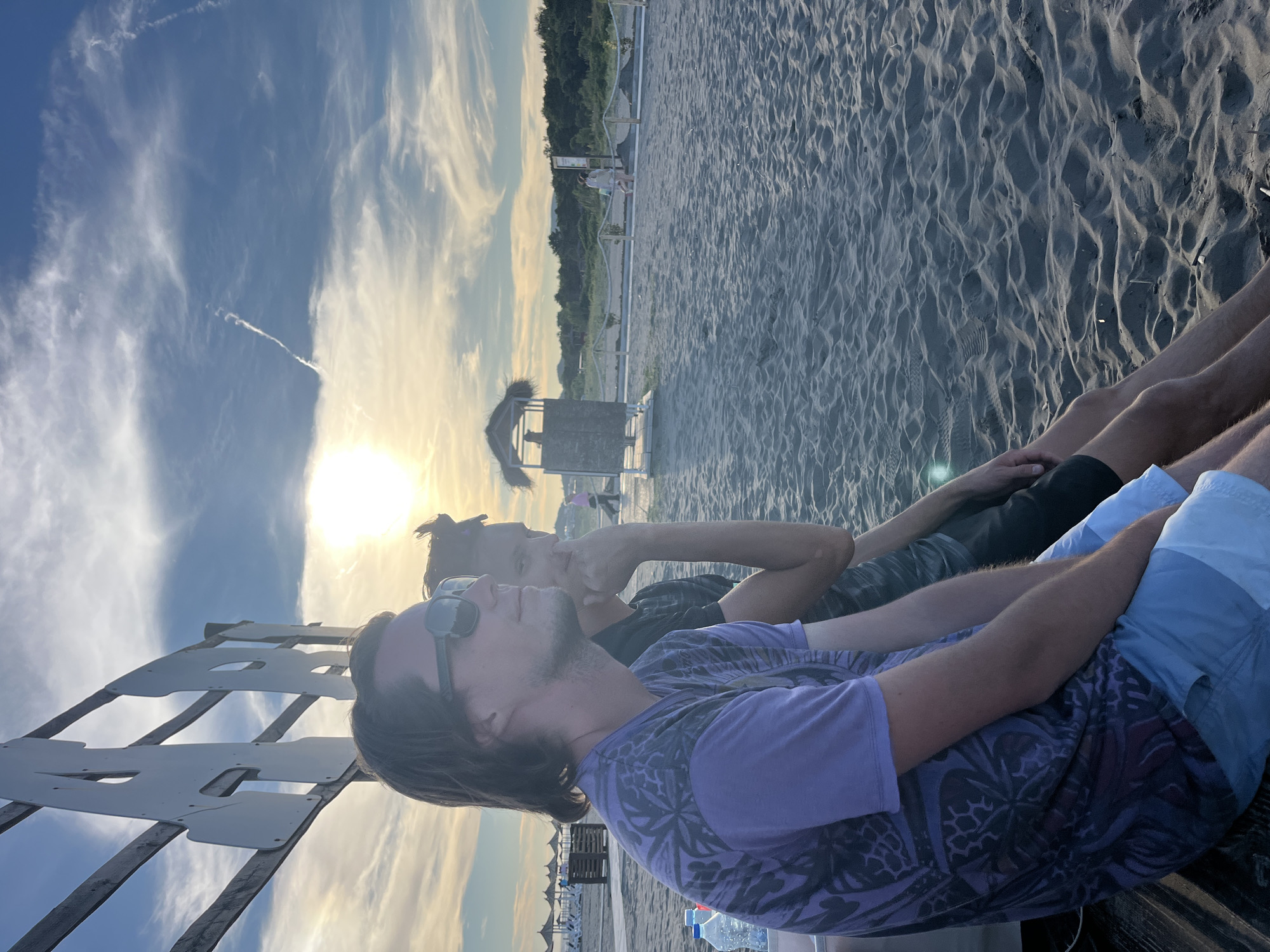 The image shows two men sitting on a wooden bench on a sandy beach. The man in the foreground is wearing sunglasses, a purple t-shirt with a black pattern, and light blue shorts. The man behind him is wearing a dark t-shirt and dark shorts. They are looking out towards the sea. In the background, there are beach umbrellas, a lifeguard tower, and vegetation. The sun is setting, casting a warm glow on the scene. The sky is partly cloudy with a contrail visible. The location is identified as the city of Pistula, Montenegro.