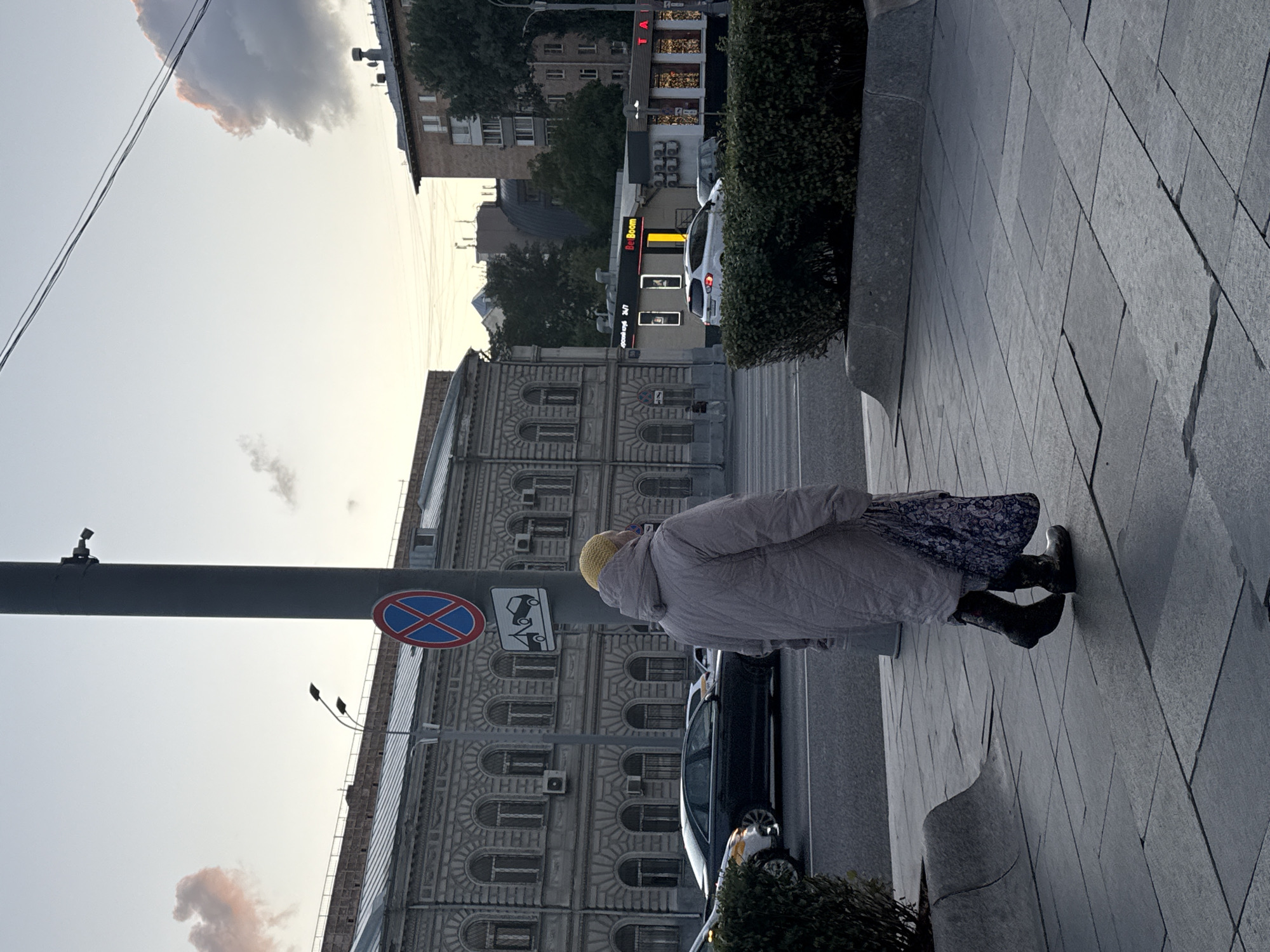 A woman wearing a light-colored puffer jacket, a floral print skirt, and dark boots walks on a paved sidewalk in Moscow, Russia. She has a yellow head covering and carries a patterned bag. In the background, a gray building with arched windows and a modern apartment building are visible. A traffic light pole with a "no parking" sign and a tow-away sign stands on the sidewalk. Cars are driving on the street in front of the buildings. The sky is a pale blue with some pink and gray clouds.