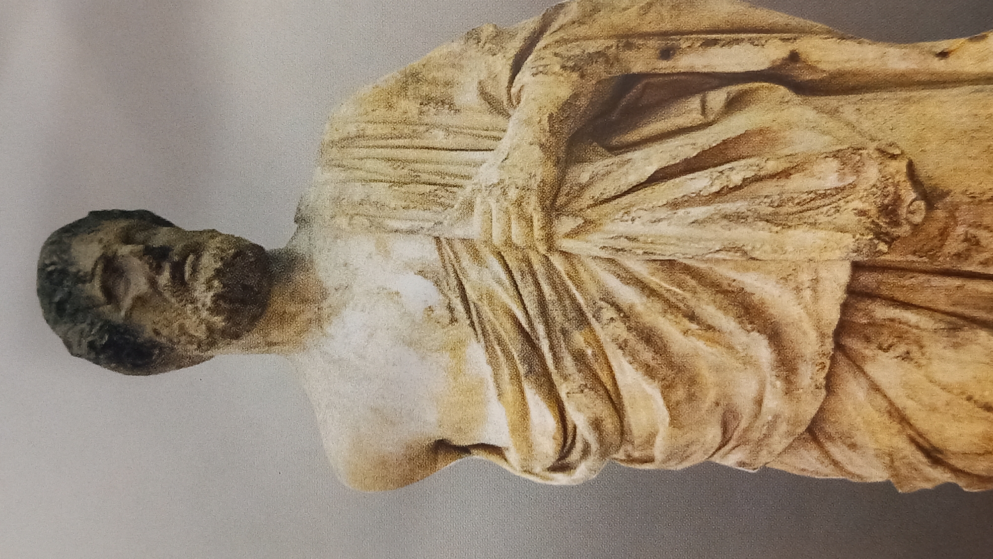 This is a full-body, waist-up, front-facing photograph of a marble statue of a bearded man, likely a philosopher or historical figure, located at the Asklepion archaeological site on the island of Kos, Greece. The statue is an ancient Hellenistic or Roman-era sculpture, characterized by its weathered and chipped surface, with a light beige and brown patina.
The subject has a stern expression, with deep-set eyes and a prominent nose. His hair is short and curly, and he has a full, tousled beard. He is draped in a classical toga, with intricate folds and pleats emphasizing the texture of the fabric. One arm is bent, and his hand is partially visible, holding the fabric of the toga. The other arm is mostly obscured by the drapery. The background is a plain, muted gray wall, likely a museum or exhibition space, with a subtle textured pattern. The lighting is even, highlighting the details of the sculpture.