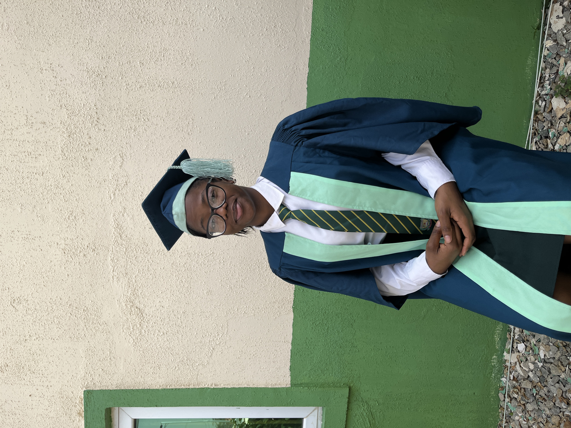 A young person is standing outdoors in Jos, Nigeria. They are wearing a graduation cap and gown in dark blue and mint green, over a white shirt and striped tie. The person is also wearing glasses. The background consists of a wall painted in two vertical stripes of beige and green and a small window with white trim is in the bottom left of the image. To the right of the green wall is an area covered in small rocks and debris. The image appears to be rotated counter-clockwise.