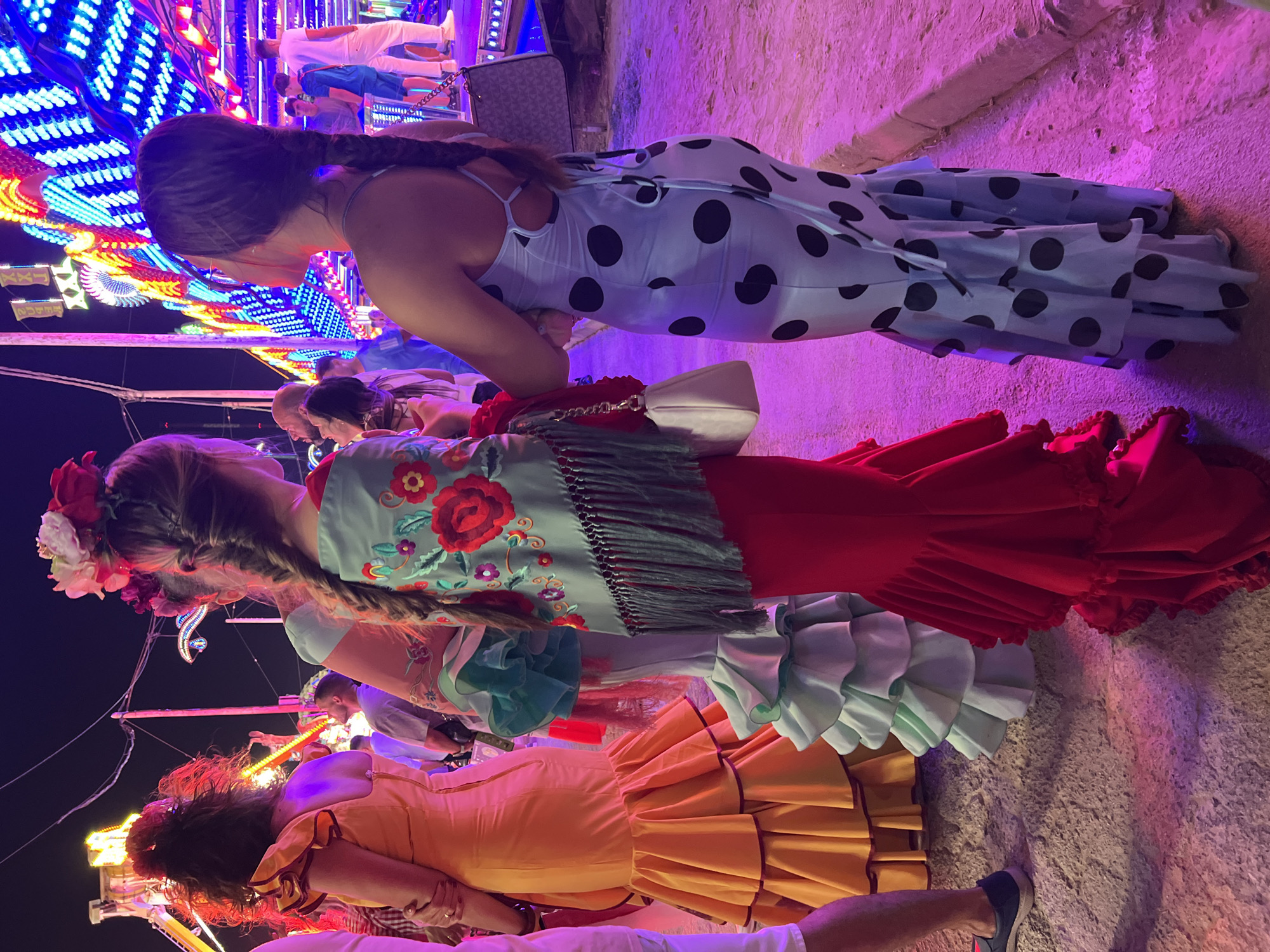 The image depicts a nighttime scene at what appears to be a fair or festival, with bright, colorful lights from amusement rides in the background. In the foreground, three women are the central focus, each wearing a distinct, voluminous flamenco-style dress.
The woman on the left is wearing an orange dress with ruffled layers and dark trim. She has her back to the camera. Next to her, a woman in a light blue and mint green layered dress also faces away. She is wearing a shawl draped over her shoulders adorned with embroidered red flowers and green foliage, with a fringed hem. She holds a white handbag.
To the right, a third woman, seen from the back and right side, is dressed in a light blue gown with large black polka dots and layered ruffles at the hem. She has her hair in a braid and is carrying a dark patterned handbag.
Several other people are visible in the background, some standing and looking at the rides, others walking. The ground appears to be paved with stone or concrete, and there is evidence of artificial lighting casting colorful hues, particularly pink and purple, on the scene. The overall atmosphere suggests a lively outdoor event. The mention of Gibraltar, Gibraltar as the location implies this event is taking place there.