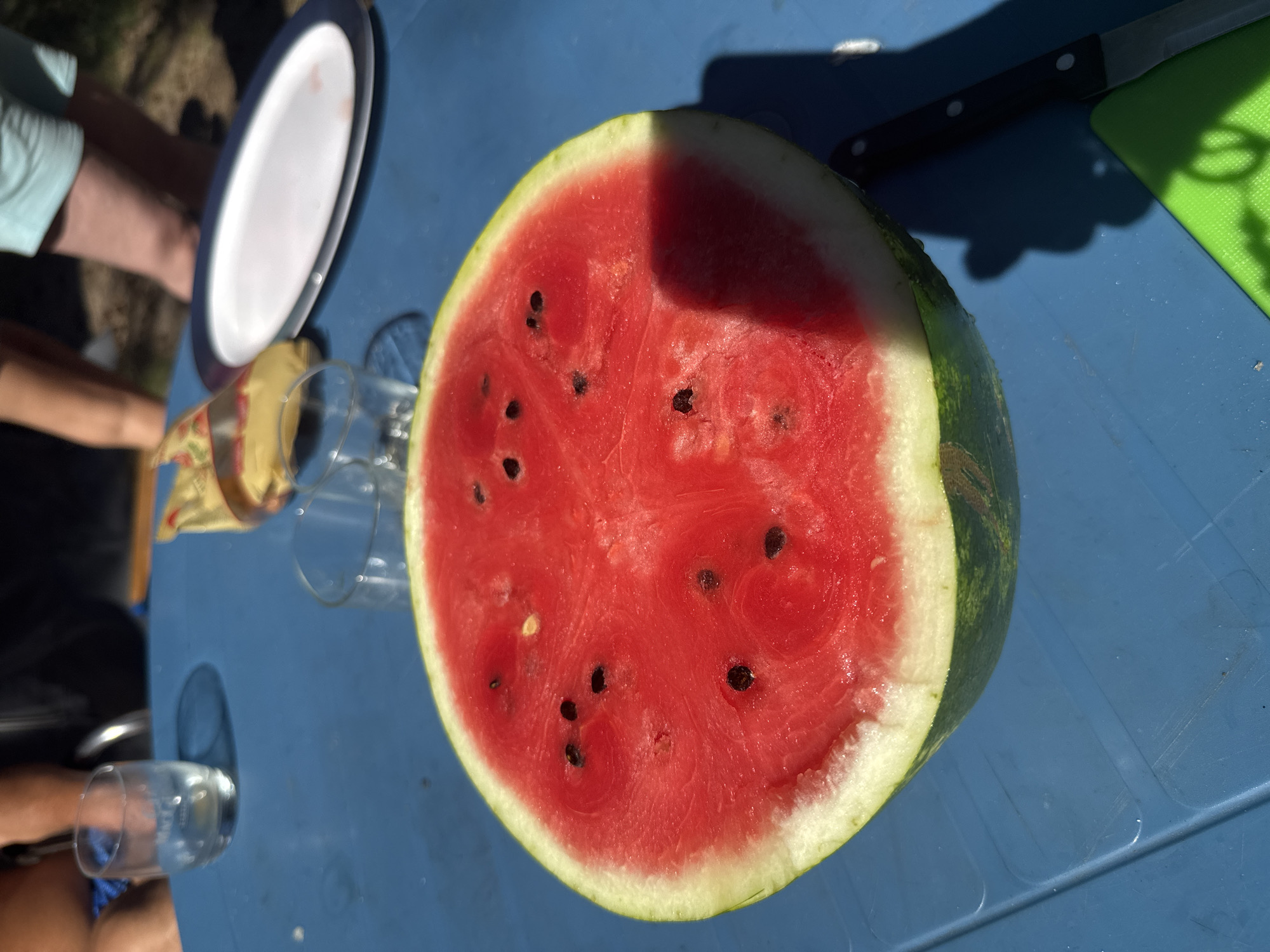 A half-eaten watermelon sits on a blue table, casting a shadow in the sunlight. Several glasses and an empty plate are also visible on the table, along with a knife and a green cutting board. In the background, a person's legs are visible, suggesting an outdoor gathering. The scene appears to be set in Zelenaya Roshcha, Russia, indicated by the provided location.