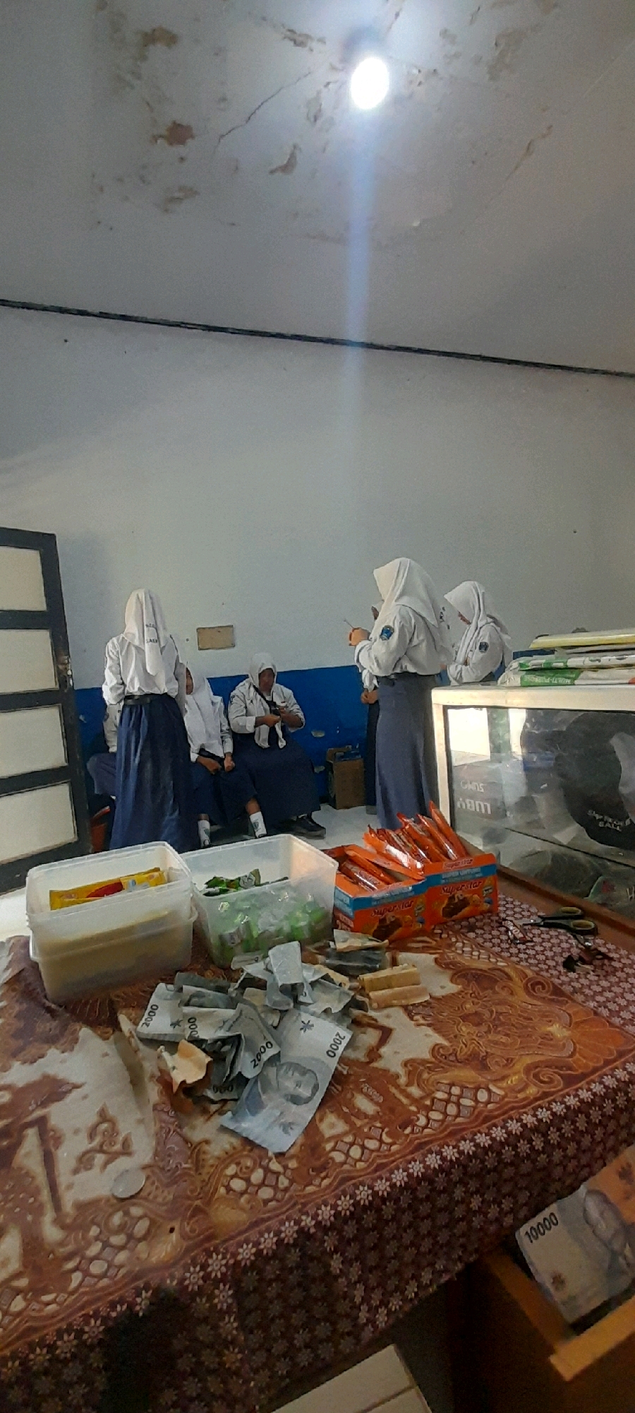 The image depicts an indoor scene that appears to be a classroom or a common area within a school in Tahunan, Indonesia. Several students, all wearing school uniforms consisting of white shirts, headscarves, and dark skirts or pants, are present. Some students are seated on a blue bench against a white wall, while others are standing.
In the foreground, a table or surface is covered with a patterned cloth. On this surface, there are various items: scattered Indonesian currency notes (including denominations of 2000 and 10000), two clear plastic containers filled with items, a box of what appear to be snacks or candy bars, and a pair of scissors. One container holds packaged items, possibly food or stationery, while the other contains green wrappers. The currency is scattered haphazardly, with some notes folded and others spread out. The snack box is labeled "SuperStar."
The environment shows signs of wear, with discoloration and cracks visible on the ceiling, and a visible light fixture. A dark door frame is present on the left side of the image. The overall impression is a candid snapshot of a casual gathering, possibly during a break or after school activities, with an emphasis on collected items and money.