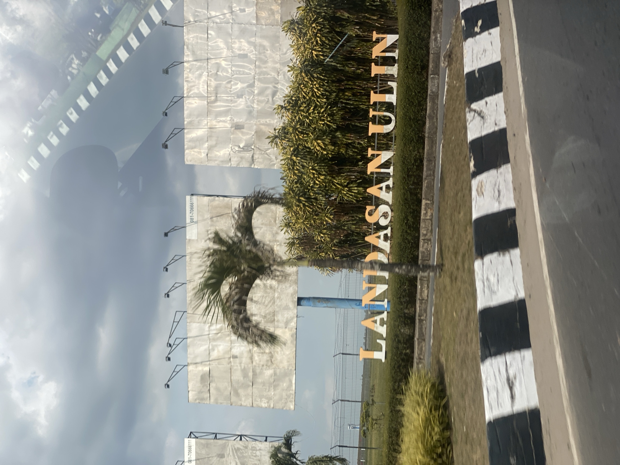 The image captures an outdoor scene with a prominent sign that reads "LANDASAN ULIN". The sign is composed of large, three-dimensional letters, some of which appear to be orange and white. Behind the sign, there is a dense line of green and yellow foliage. Further in the background, three large, blank billboards are visible, mounted on poles. The billboards are covered in a light-colored, textured material. The sky above is cloudy with varying shades of gray. In the foreground, a road with a black and white striped barrier is visible, suggesting a traffic or roadside setting. The overall impression is a roadside entrance or landmark in a somewhat overcast environment. The location is identified as Landasan Ulin, Banjarbaru, Indonesia, which is known for its airport and a significant military airbase.