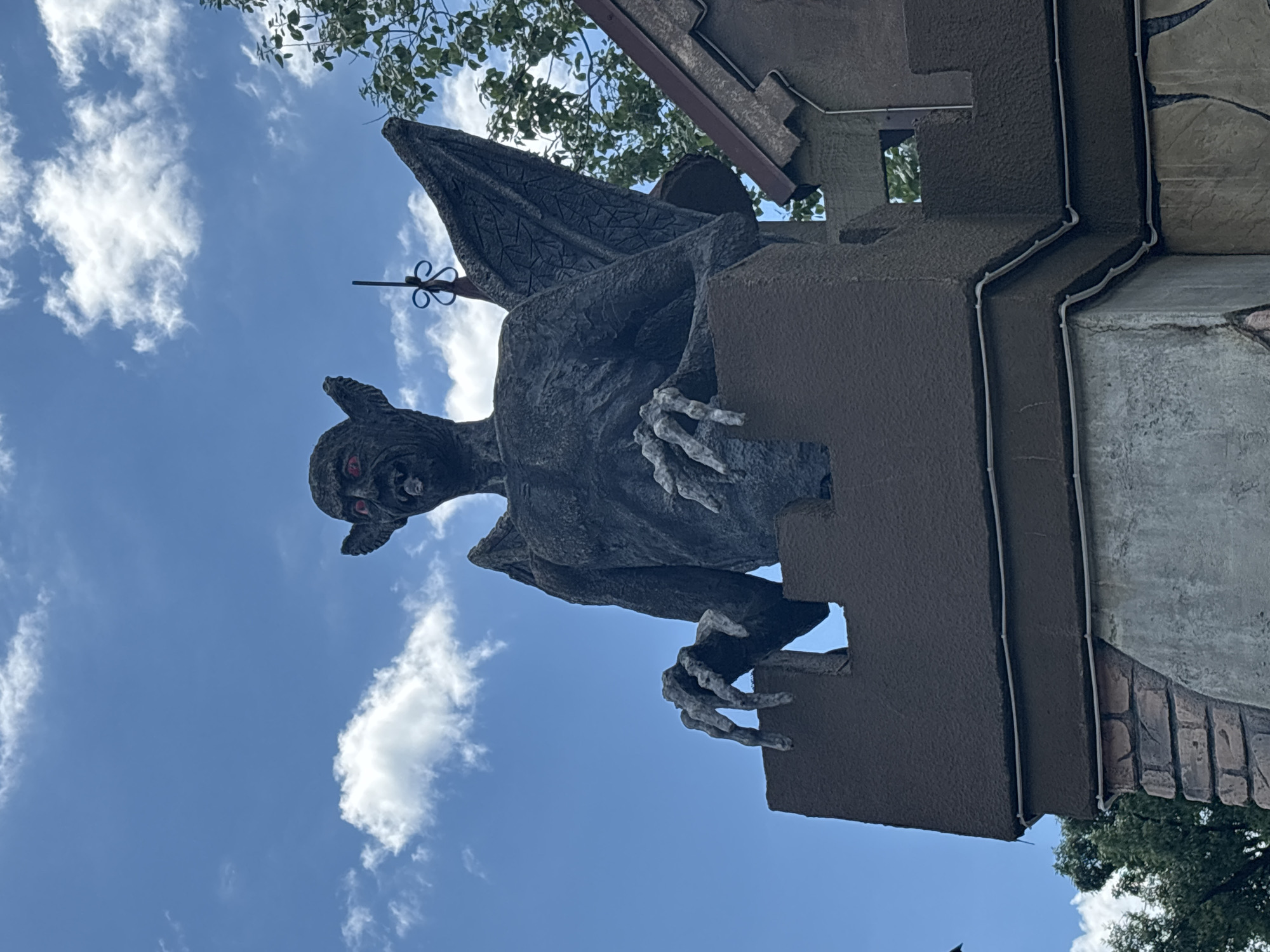A grotesque, dark gray gargoyle statue with red eyes and a protruding tongue is perched atop a dark brown, castle-like structure. The gargoyle's clawed hands rest on the parapet. Its textured, leathery wings are spread behind it. The statue is situated against a bright blue sky with scattered white clouds. To the right of the gargoyle, a portion of a brown roof with a jagged edge is visible, along with green foliage. The lower part of the structure appears to be made of brick and a rough concrete-like material. The scene is set in the city of Sochi, Russia.