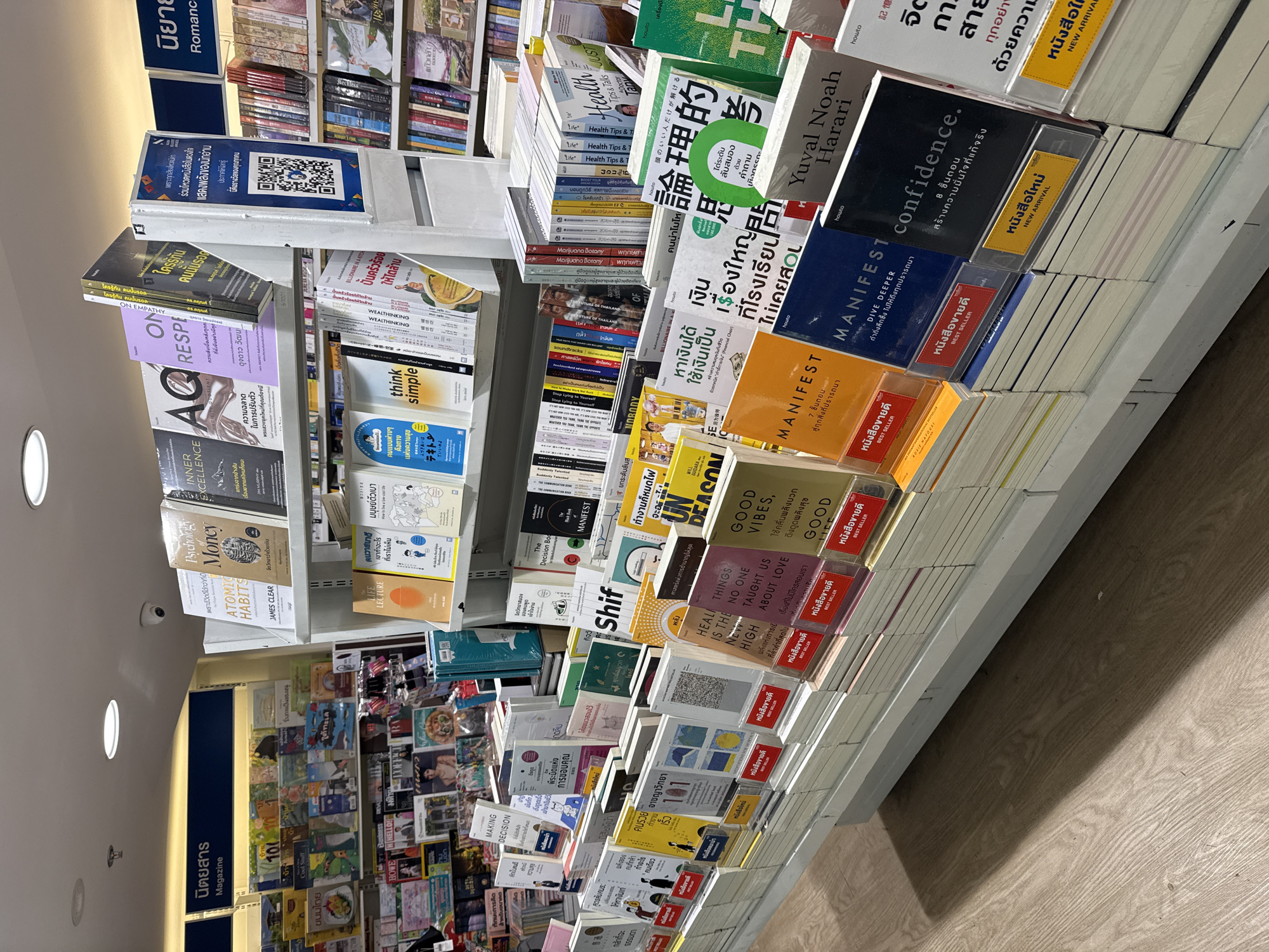 The image depicts a bookstore interior, likely a branch of Asia Books, located in the city of Nong Prue Subdistrict, Thailand. The store features brightly lit shelves stocked with a diverse selection of books, primarily in English and Thai, covering genres such as self-help, business, and general interest. A prominent display showcases "Best Seller" and "New Arrival" labels, indicating a focus on popular and recently released titles. Among the visible books are "ATOMIC HABITS," "THE LIFE LECTURE," "MANIFES T," and "Confidence." QR codes are also visible on display stands, suggesting promotional activities. The environment is clean and organized, with wooden flooring and overhead lighting contributing to a retail atmosphere. There are no individuals present in the frame.