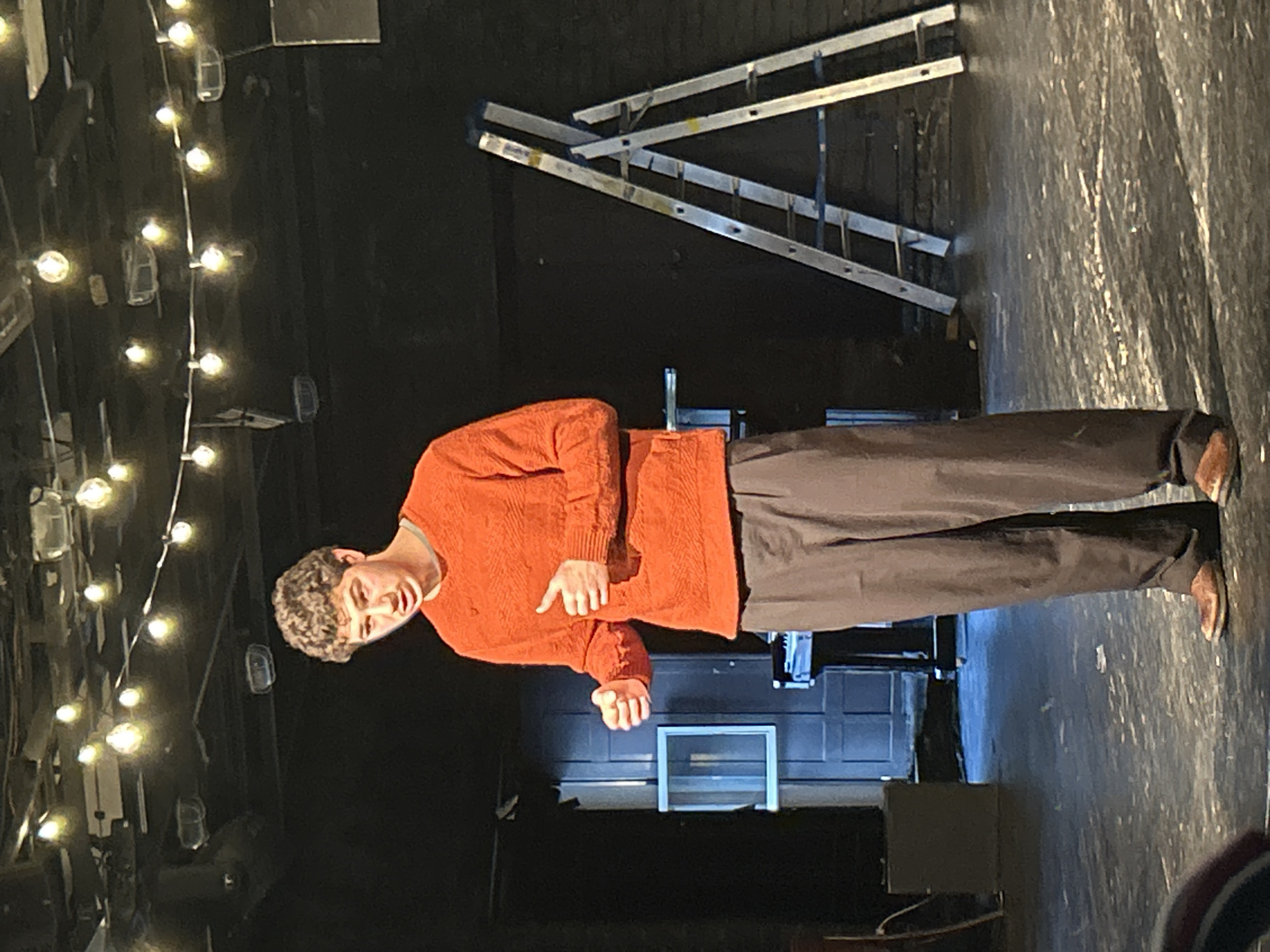 An indoor scene depicts a young man standing on what appears to be a stage or rehearsal area. He is positioned slightly off-center to the left, facing towards the viewer with a thoughtful or engaged expression, mouth slightly open as if speaking or singing. He has short, curly dark hair and is wearing a textured, orange-red long-sleeved sweater, dark grey or brown trousers, and brown dress shoes. His hands are slightly raised and open, suggesting a gesture or moment of pause.
To the man's left, a string of clear-bulbed lights hangs vertically, providing a warm glow. The background is predominantly dark, indicating a low-light environment typical of a stage. In the upper right corner, a silver-colored metal ladder is visible, seemingly propped against a wall or backdrop. Below and behind the man, indistinct dark shapes and structures can be seen, including what looks like a dark, panelled door and a white, rectangular frame object on the floor. The stage floor is dark and appears slightly worn or scuffed. No visible text is present in the image. The overall atmosphere suggests a performance, rehearsal, or production setting.