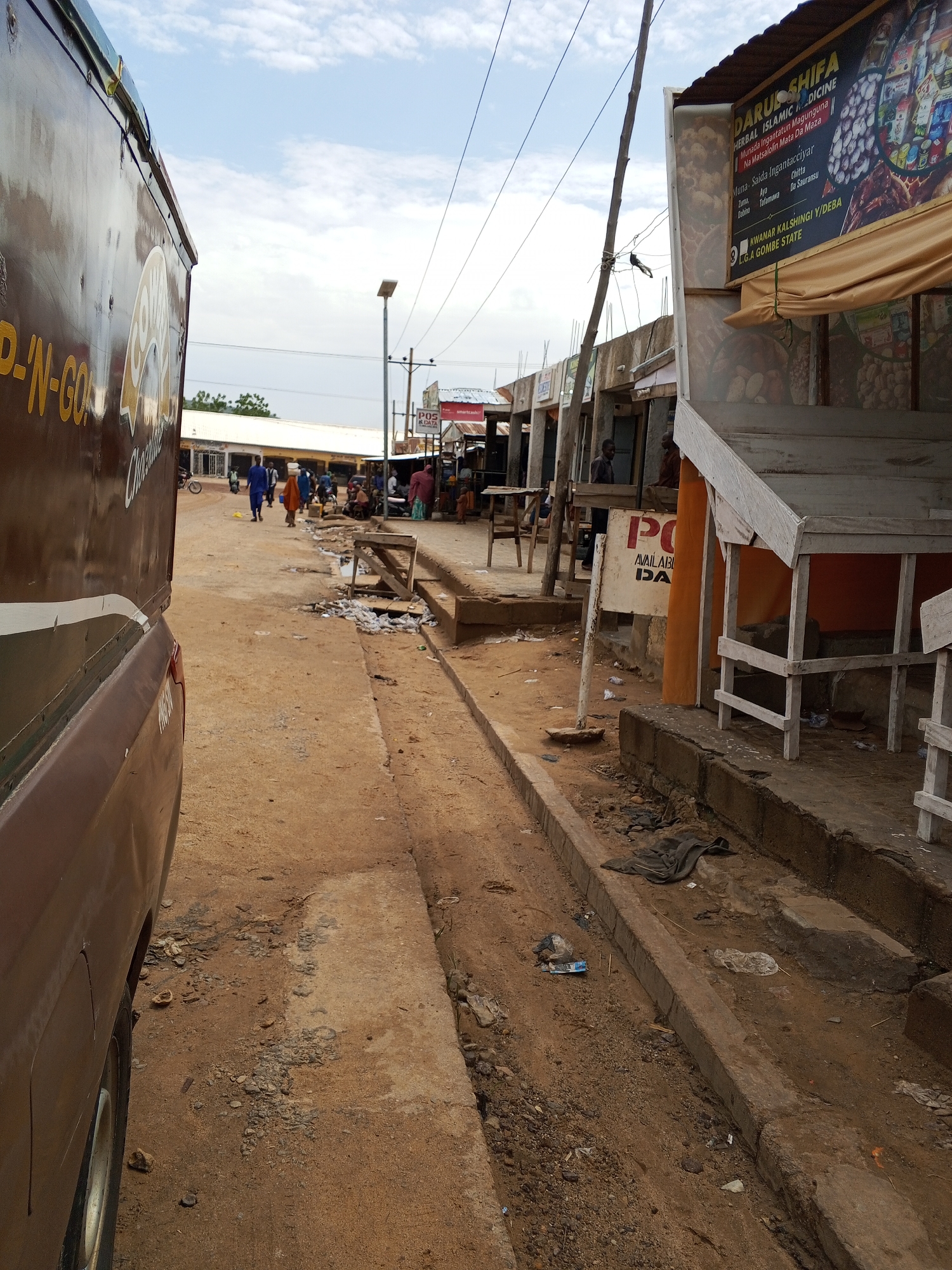 This is a ground-level, full-shot photograph taken on a dusty street in Galadima, Nigeria. The image captures a street scene with several elements, including a parked brown van on the left, a row of market stalls or shops on the right, and people walking and standing.
The van on the left is a brown, boxy vehicle with text partially visible on its side. The text "N-GO" is clearly legible, suggesting it might be a delivery or transport vehicle. The van occupies a significant portion of the left side of the frame.
To the right of the van, the street is unpaved and dusty, with patches of gravel and debris. A concrete drainage channel runs along the edge of the pavement in the foreground.
The right side of the image is dominated by a row of market stalls. One stall has a prominent sign with "DARU SHIFA HERBAL ISLAMIC MEDICINE" written on it, along with text in what appears to be Arabic or a similar script. The stall itself has wooden structures and is covered by a beige awning. Another stall has a sign with "POS & DATA" and a small satellite dish visible. These stalls are open-fronted, revealing goods or displays within.
Several people are present in the scene. Some are walking down the street, including individuals carrying items on their heads. Others are standing near the market stalls. Two men are visible near one of the stalls, one wearing a dark shirt and the other in a brown garment. A child is also visible near the stalls.
The environment appears to be a busy marketplace in an urban or semi-urban setting. The sky above is partly cloudy, indicating daylight. The overall impression is one of everyday activity in a Nigerian town.