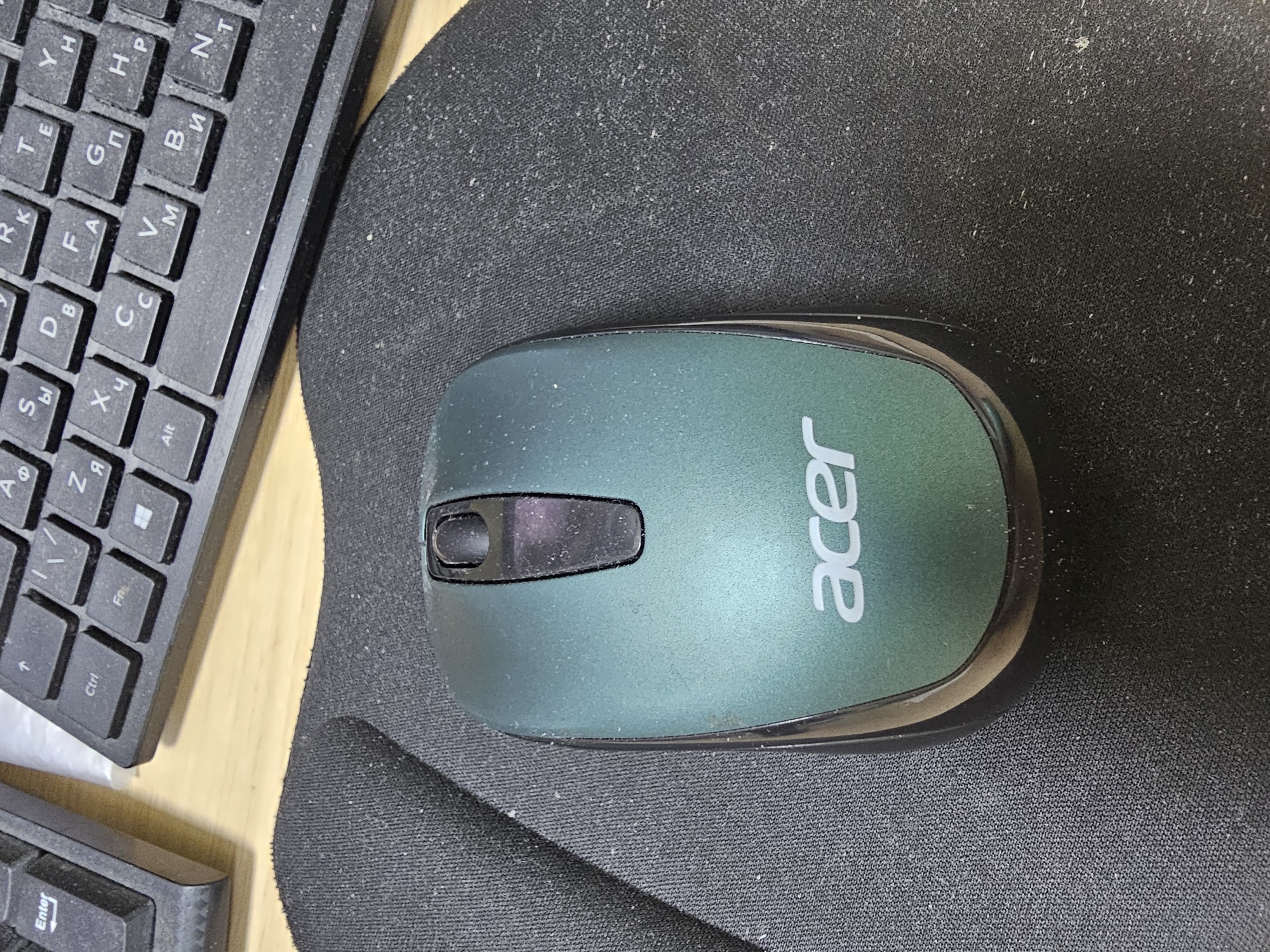 A close-up, top-down shot shows a dusty, teal-colored Acer computer mouse resting on a black mousepad. To the left of the mousepad, a portion of a black keyboard is visible. The keyboard has some Cyrillic lettering on its keys, and dust is also apparent on the keys. The scene appears to be an indoor workspace. No specific location in Saint Petersburg, Russia is indicated by the image content.