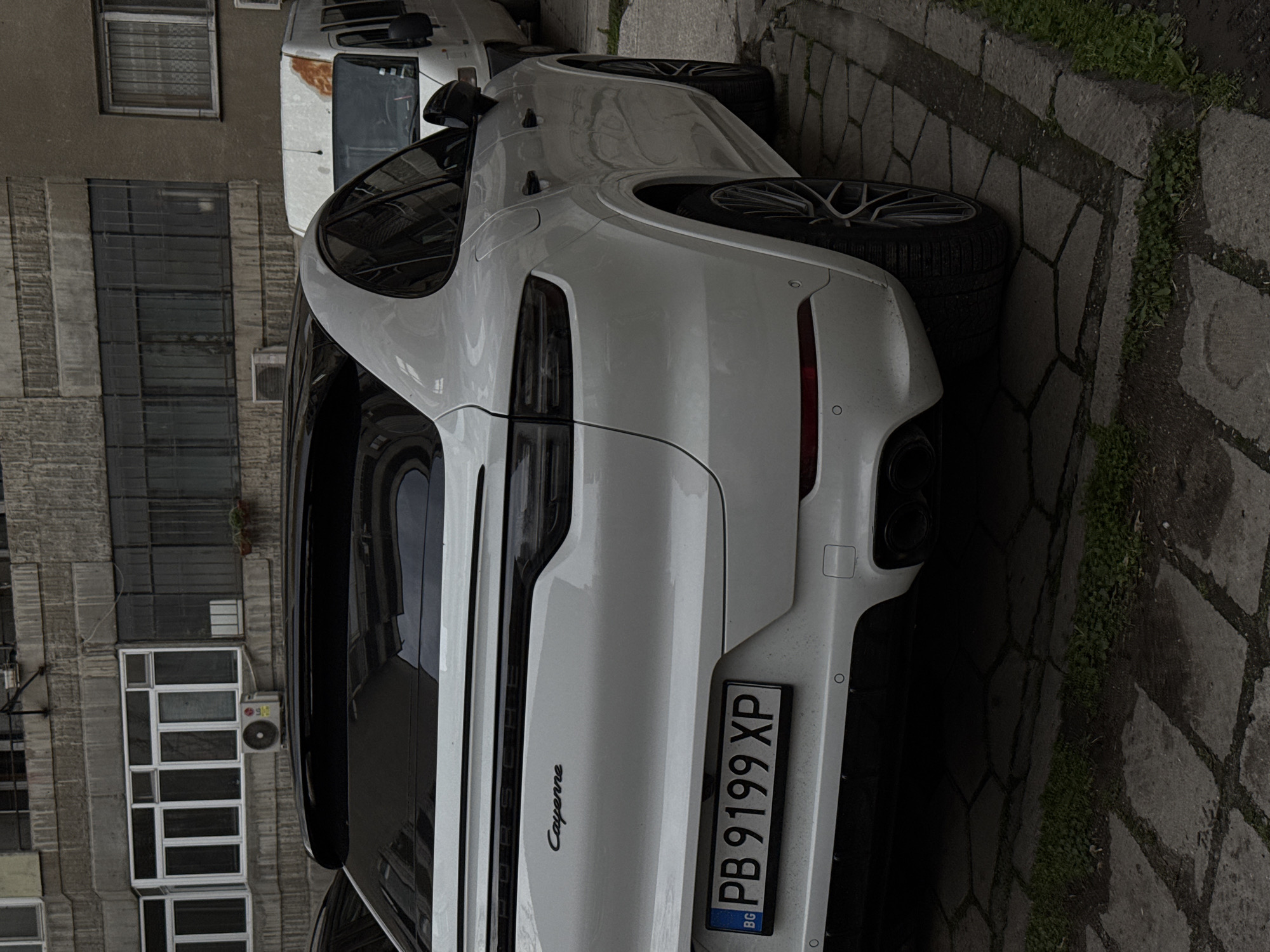 The image shows the rear of a white Porsche Cayenne parked on a paved street in an urban environment. The car's Bulgarian license plate reads "PB 9199 XP." Behind the car, there is a building with windows and an air conditioning unit. A white van is partially visible in the background. The street is paved with interlocking stones, and some vegetation grows along the edge. The image does not provide enough information to confirm the specific location in Plovdiv.