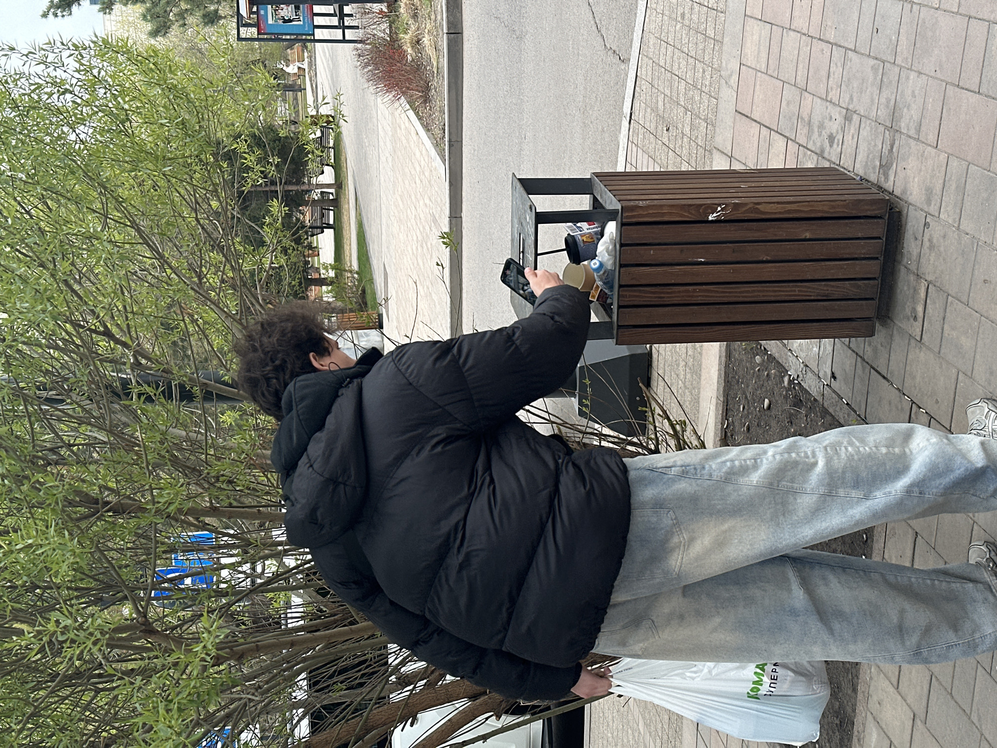 A person with dark, curly hair is discarding waste into a public trash can with a mobile phone in hand. The person wears a black puffer jacket, light-wash denim jeans, and grey athletic shoes. They hold a white plastic bag in their left hand. The scene appears to be set on a sidewalk near a tree in Krasnoyarsk, Russia.