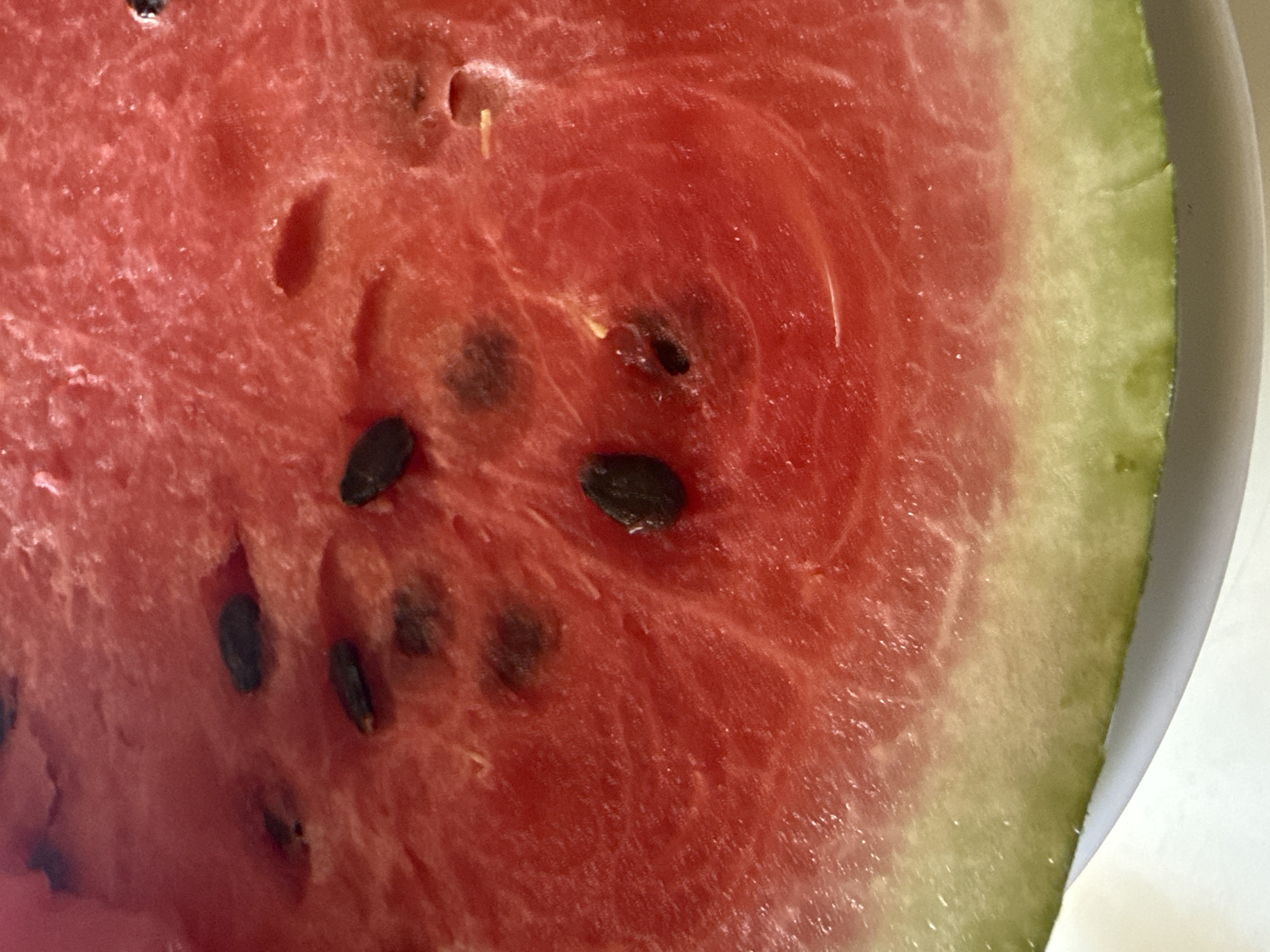A close-up, overhead shot shows a segment of a ripe watermelon slice. The vibrant pink flesh is dotted with dark, oval seeds, some of which are partially submerged in the juicy pulp. The exterior rind transitions from a pale green to a darker green, and appears slightly textured. The slice rests on a white surface, likely a plate.