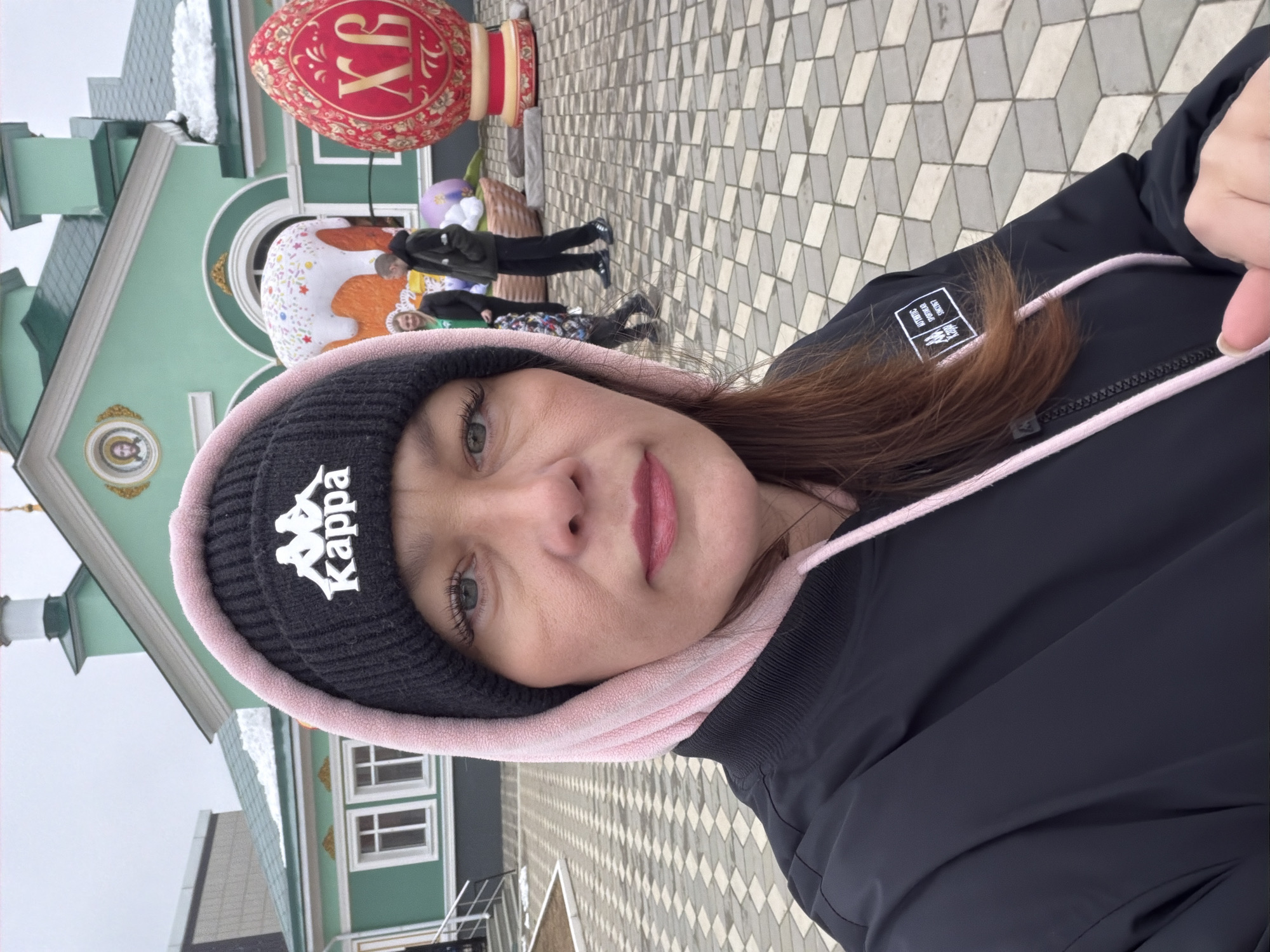 A selfie of a woman wearing a black Kappa beanie and a black jacket with a pink hood lining is taken in front of an elaborate building and Easter-themed decorations. The building is painted green and white, with architectural details including windows and what appears to be an icon set into the facade. Behind the woman, two large, colorful Easter eggs are visible: one is red with Cyrillic script "XB", and the other is decorated like a traditional Easter cake with white frosting and colorful sprinkles.
In the background, there are at least two individuals walking on a paved area composed of interlocking gray and white geometric tiles. The scene is overcast.
Based on the visual cues and the knowledge that such Easter decorations are commonly found in public spaces during the Easter season, the location is likely Chaikovsky, Russia, during the period of Orthodox Easter celebrations, with the decorations set up in a public area near an Orthodox church.