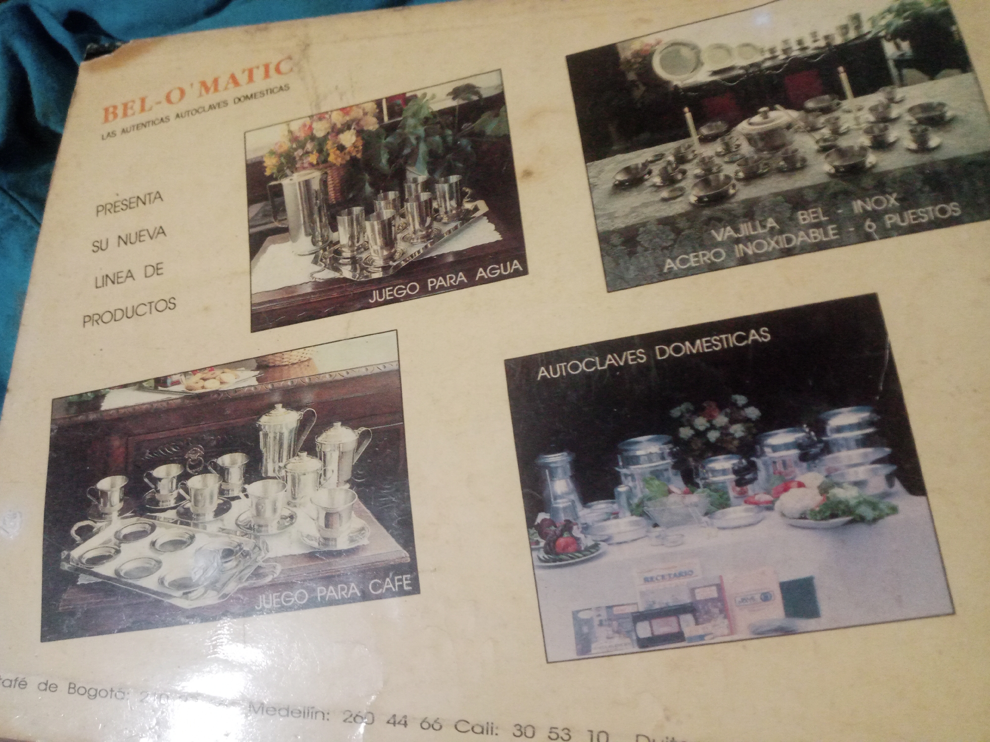 The image is a photograph of a brochure or advertisement for "BEL-O'MATIC LAS AUTENTICAS AUTOCLAVES DOMESTICAS." The brochure features several product images.
The top left section displays text that reads "PRESENTA SU NUEVA LINEA DE PRODUCTOS." Below this, there is a photograph of a silver pitcher and several glasses on a tray, with text that says "JUEGO PARA AGUA."
To the right of this is a photograph of a table setting with a tea set, including a teapot, cups, and saucers. Text below this reads "VAJILLA BEL - INOX ACERO INOXIDABLE - 6 PUESTOS."
In the bottom left is another photograph showing a silver coffee set with a tray, cups, and pots, with text below that says "JUEGO PARA CAFE."
The bottom right of the brochure shows a collection of stainless steel kitchenware, including pots and serving dishes, arranged on a table. Some vegetables and fruits are visible on a platter. Text at the top of this section reads "AUTOCLAVES DOMESTICAS."
The brochure appears to be printed on a light-colored paper with some age-related discoloration. At the very bottom, there is some faint text with city names and numbers, suggesting contact information or distribution points. One line of this text is partially visible and reads "Bogotá: 210" and "Medellín: 260 44 66 Cali: 30 53 10". The overall context of the image suggests a product advertisement for kitchen appliances and tableware.
There is no information in the image that provides the location in the city of Santa Ana, Venezuela.