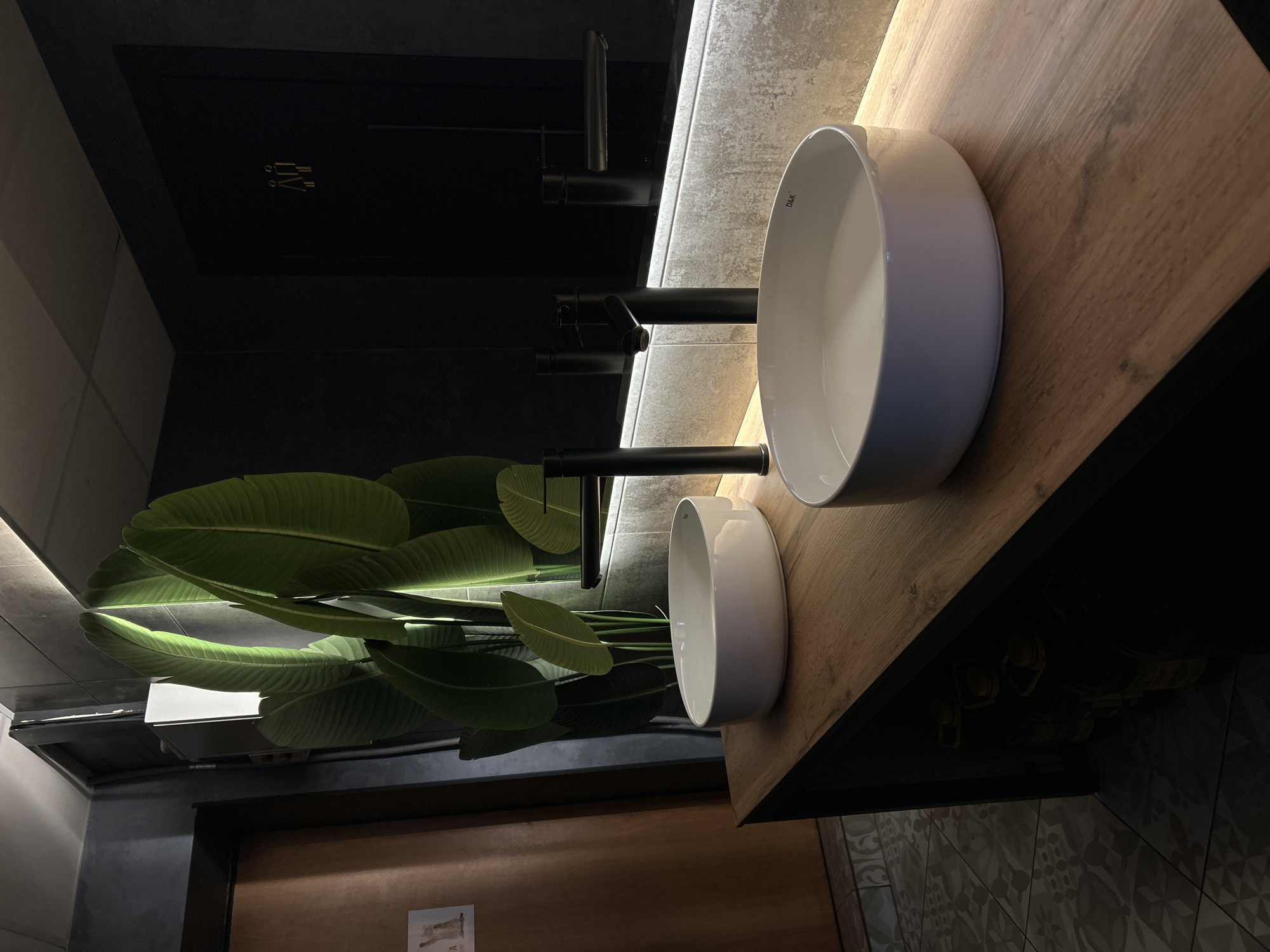 The image shows a bathroom interior with a focus on two white, circular vessel sinks placed on a wooden countertop. Black, minimalist faucets are positioned above each sink. A large, green leafy plant is situated near the sinks, adding a touch of nature to the space. The background includes a dark-colored wall and a mirror reflecting the scene. The tile floor has a geometric pattern. The location is Chaikovsky, Russia.