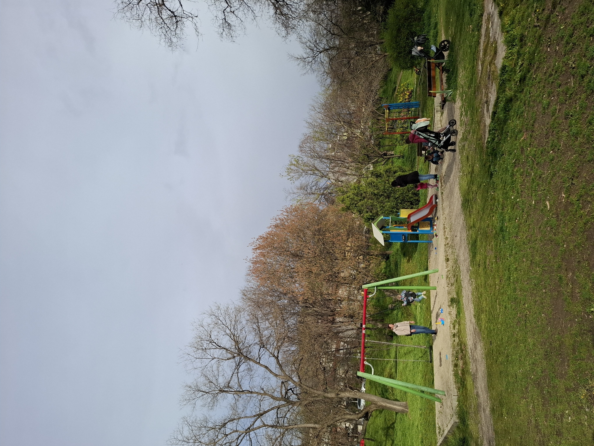 The image shows a park in Plovdiv, Bulgaria. The location is likely a children's playground. There is a gravel path running diagonally across the frame, separating a grassy area. Several playground structures are visible, including swings, a slide, and other climbing frames. Several people are present, including adults and children. Some individuals are using the playground equipment, such as the swing. The weather appears overcast, with a cloudy sky. The trees are bare or have budding leaves, suggesting it is either early spring or late autumn.