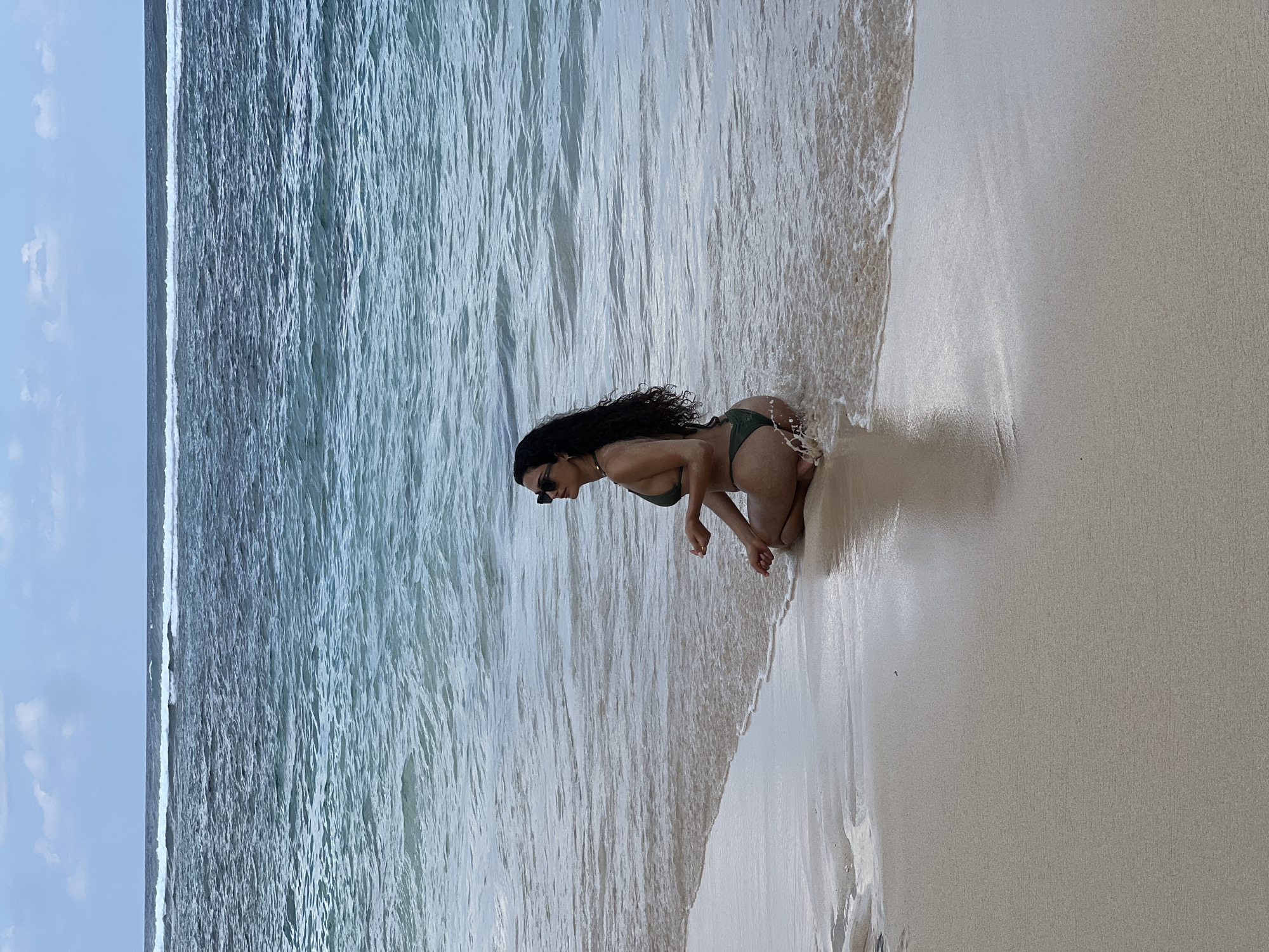 The rotated image captures a tropical beach scene, with the horizon appearing vertically on the left side. The main subject is a woman with long, dark, curly hair, dressed in a dark green bikini and sunglasses. She is kneeling or crouching gracefully in the shallow water at the very edge of the ocean, where gentle waves meet the shore. Water splashes visibly around her as she interacts with the incoming tide.
The setting features a wide expanse of clear, blue-green ocean water, characterized by subtle ripples and small waves. This transitions smoothly into a light-colored, almost white, sandy beach. The wet sand near the water's edge reflects the light, indicating the presence of a receding wave. In the background, a light blue sky with faint white clouds suggests a bright, daytime setting, likely sunny or partly cloudy. The overall mood is one of tranquility and enjoyment of nature. There is no visible text in the image. The scene is consistent with the beautiful coastal environments found in Kuta Selatan, Indonesia.