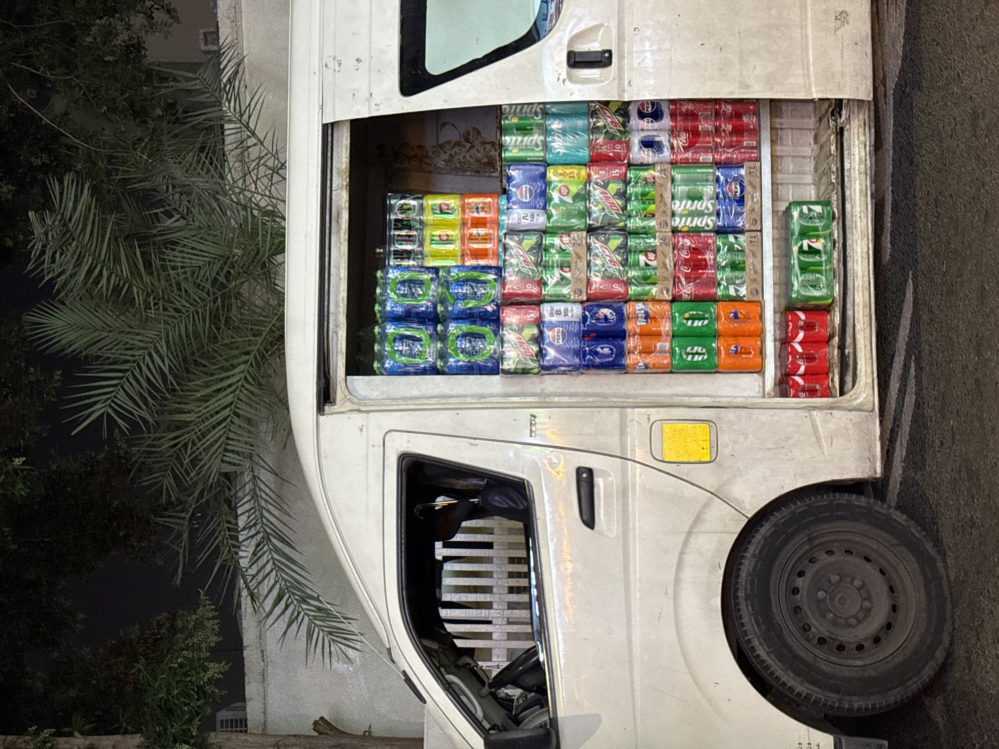 A white truck is parked with its rear cargo area open, revealing a variety of canned beverages neatly stacked inside. The beverages include Pepsi, Coca-Cola, Sprite, Mountain Dew, and Fanta. The truck is parked on a paved surface, likely a road or parking lot. A palm tree is visible to the left of the truck. The location is likely Dubai, United Arab Emirates, based on the types of beverages typically sold in the region and the architectural style in the surrounding environment. The scene appears to be at night, as indicated by the darkness and the presence of artificial lighting. There are no visible people in the image.