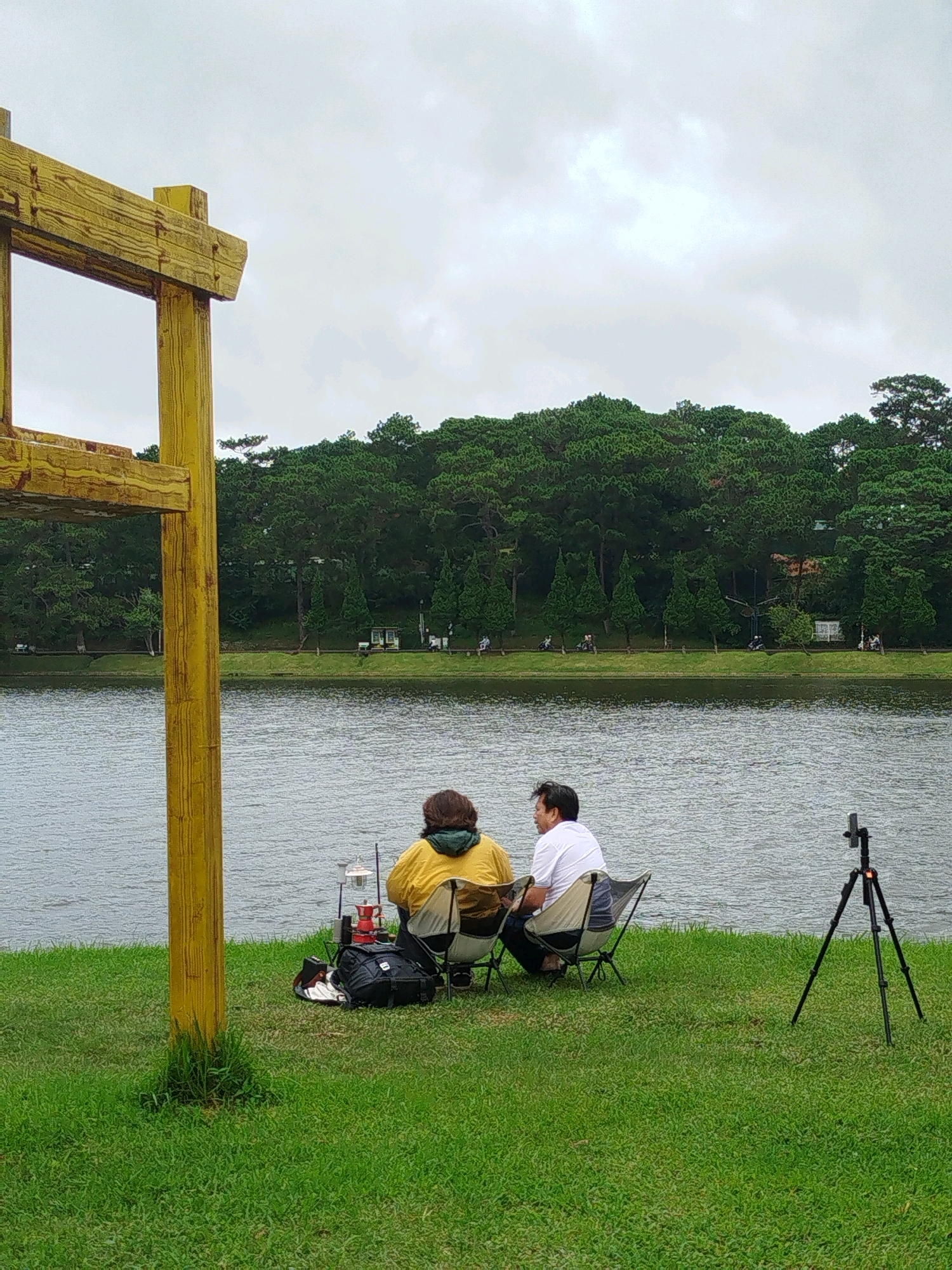 The image depicts a tranquil lakeside scene in Da Lat, Vietnam. In the foreground, a large yellow wooden structure, possibly part of a pavilion or decorative element, stands to the left. The area is covered in lush green grass, indicating a park or recreational area.
Two individuals are seated in folding chairs by the lake. The person on the left is wearing a yellow jacket and a green scarf, facing away from the camera and towards the water. To their right, a person wearing a white t-shirt is also seated, facing the lake and slightly to the side. Between the two individuals, there are camping chairs, a backpack, and what appears to be a portable stove and coffee maker setup.
The lake itself occupies the middle ground, with calm, rippling water reflecting the overcast sky. In the background, a dense line of pine trees forms a dark green border along the opposite shore. Beyond the trees, a few scattered buildings are visible, suggesting the presence of a small town or resort.
To the right of the seated individuals, a black tripod with a camera or phone mounted on it is positioned on the grass, pointing towards the lake, suggesting the possibility of photography or filming. The overall atmosphere is one of peaceful relaxation and enjoyment of nature, characteristic of Da Lat's scenic landscapes.