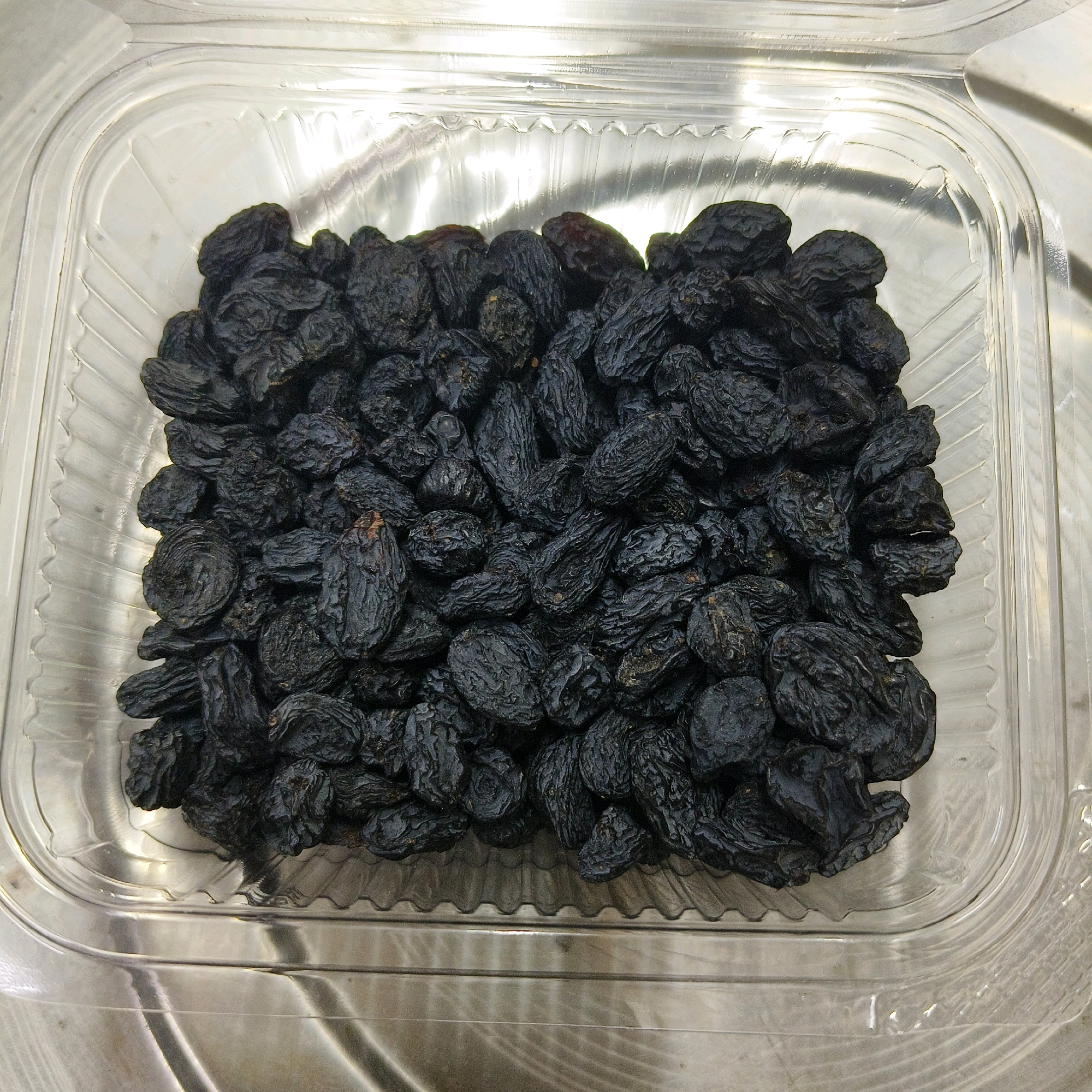 A close-up, overhead shot shows a clear plastic rectangular container filled with dark, wrinkled raisins. The container is filled to capacity, with the raisins piled high in the center. The plastic container has a ridged pattern along its base and a smooth, rounded rim. The surface the container rests on appears to be metallic and reflective, with horizontal lines and subtle light streaks. The lighting is bright, creating highlights on the plastic and the surface below. The image focuses on the texture and deep color of the raisins.