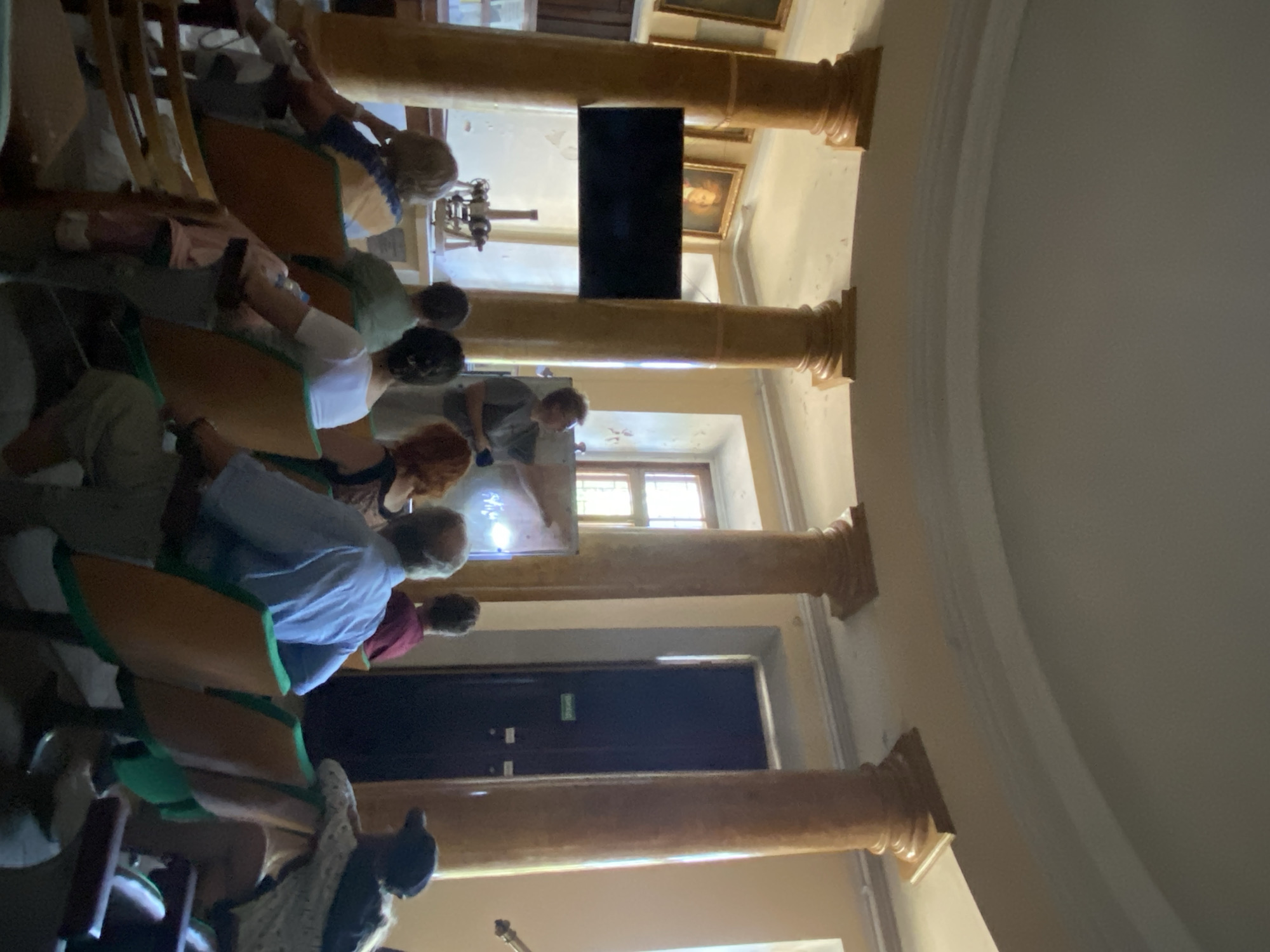 The image depicts an indoor scene with a group of people seated in chairs, facing a woman who appears to be giving a presentation. The setting has a classical feel, with large, ornate columns supporting a curved ceiling. A black flat-screen television is mounted on the wall between two of the columns. Behind the presenter, a whiteboard or projection screen is visible, with some writing on it. In the background, there are framed pictures on the walls and a door. The lighting suggests a daytime scene. The people in the audience are diverse in age and gender, and some are paying attention to the presenter while others are looking elsewhere. The overall atmosphere appears to be educational or informational.