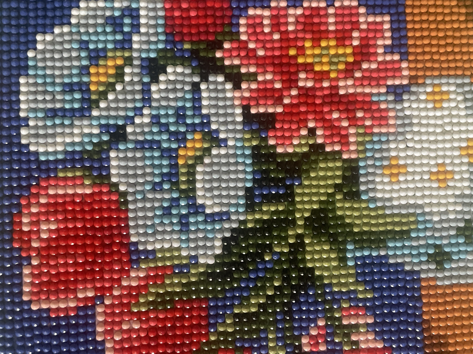 This is a close-up image of a completed diamond painting depicting a bouquet of flowers. The flowers are rendered in shades of red, white, blue, and orange with yellow accents. The background is a mix of blue and green. The image shows the texture and color transitions created by the individual diamond beads. There is no evidence that the image was taken in Saratov, Russia.