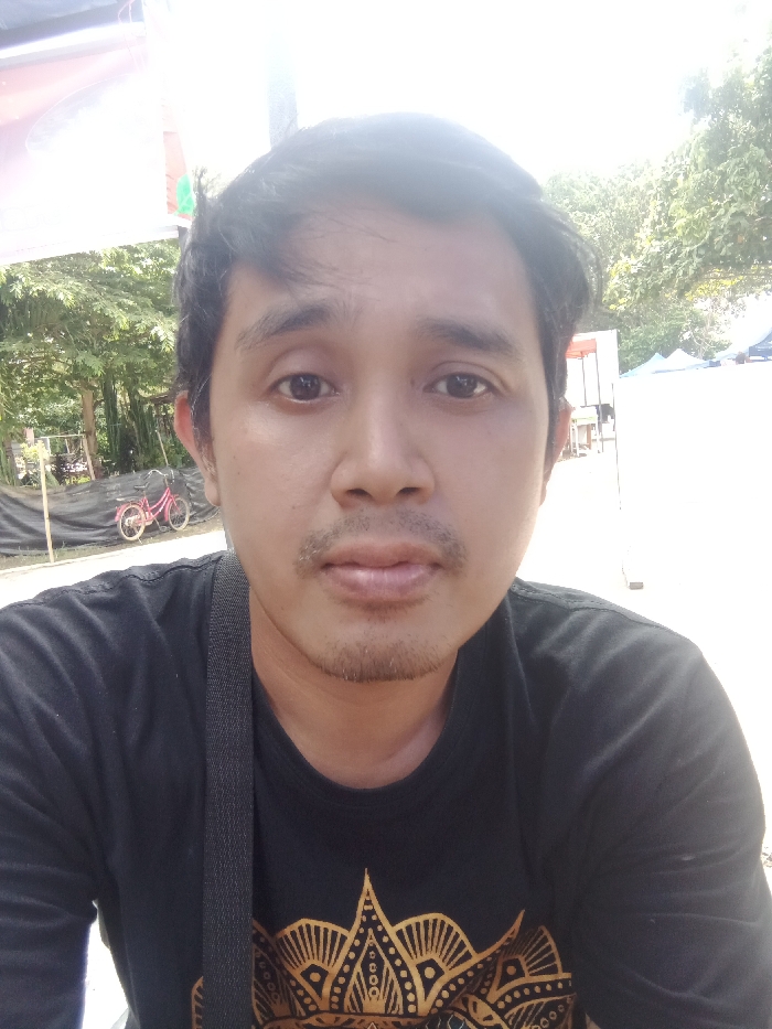 A medium-shot selfie captures a man in a black t-shirt with a golden design. The background features greenery and hints of tents. His complexion is fair, with dark hair and a thin mustache. A strap crosses his torso. The lighting appears bright, creating highlights and shadows on his face. A bicycle is visible in the distance.