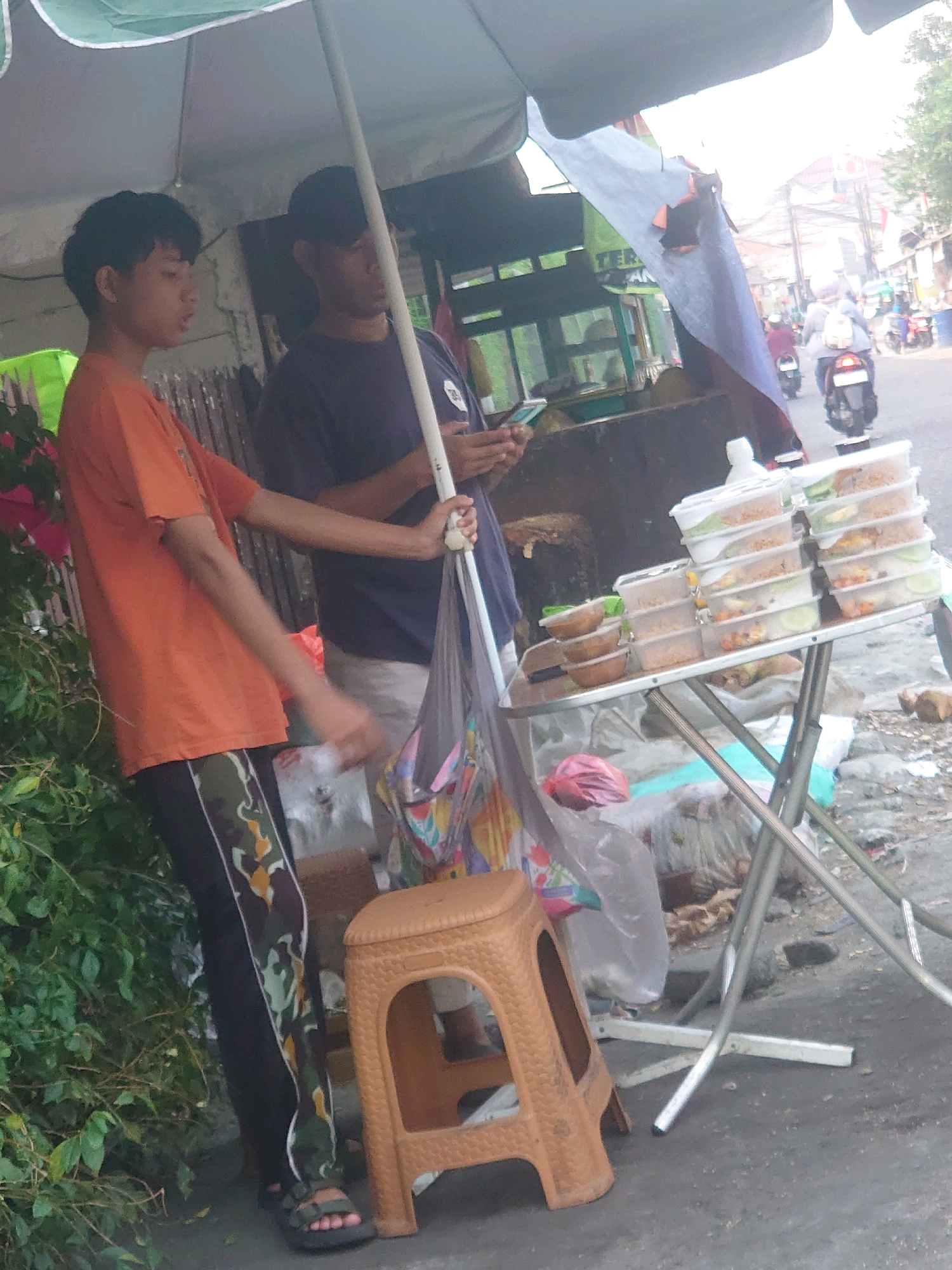 In Depok, Indonesia, two young men are seen under a green and grey umbrella, likely operating a food stall. The man on the left, wearing an orange t-shirt and camouflage pants, is standing and holding a white pole. The man on the right, in a dark t-shirt and black baseball cap, is holding a mobile phone and a white pole. In front of them, a metal folding table displays several stacks of clear plastic containers filled with food, some with cucumber slices. A brown plastic stool sits to the left of the table. Behind the table, a variety of goods are visible, including plastic bags and what appear to be plants. The scene is set along a street with visible traffic, including motorcycles, suggesting a bustling urban environment. The ground is paved and appears to be a sidewalk or roadside.