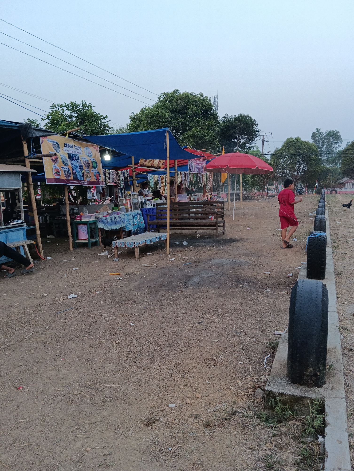 The image depicts an outdoor market scene in Pematang Jaya, Indonesia. The market consists of several stalls with blue and red awnings. One stall prominently displays a banner that reads "KEDAI EMON" and appears to advertise food and drinks. Various items are displayed for sale on tables, including what seem to be packaged goods and beverages. A red umbrella is set up near the stalls.
In the foreground, a young person in a red t-shirt and shorts is walking away from the camera. To the right of the image, a line of used tires is arranged along a concrete curb, separating the market area from a dirt and gravel ground. A chicken is visible foraging on the ground near the tires and stalls. In the background, trees, utility poles, and what looks like a telecommunications tower are visible, suggesting an urban or semi-urban environment. The ground is covered with dirt, small stones, and scattered debris. The overall lighting suggests it is late afternoon or early evening, with an overcast sky.
