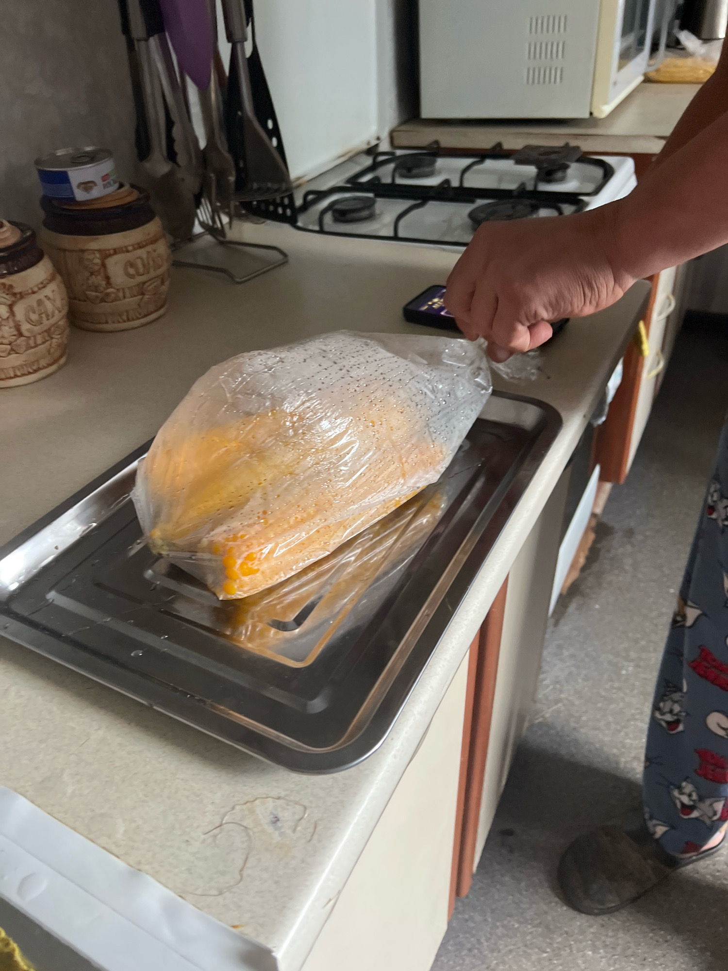 The image depicts a kitchen counter in Drohobych, Ukraine, where a person is preparing food. A large piece of yellow food, possibly a pumpkin or squash, is encased in a clear plastic bag and placed on a metal tray. A hand is visible reaching towards the bag, holding a mobile phone. The background shows kitchen appliances, including a stove and microwave, and decorative jars on a shelf. The overall scene suggests a domestic setting, possibly a home kitchen, where food preparation is taking place.