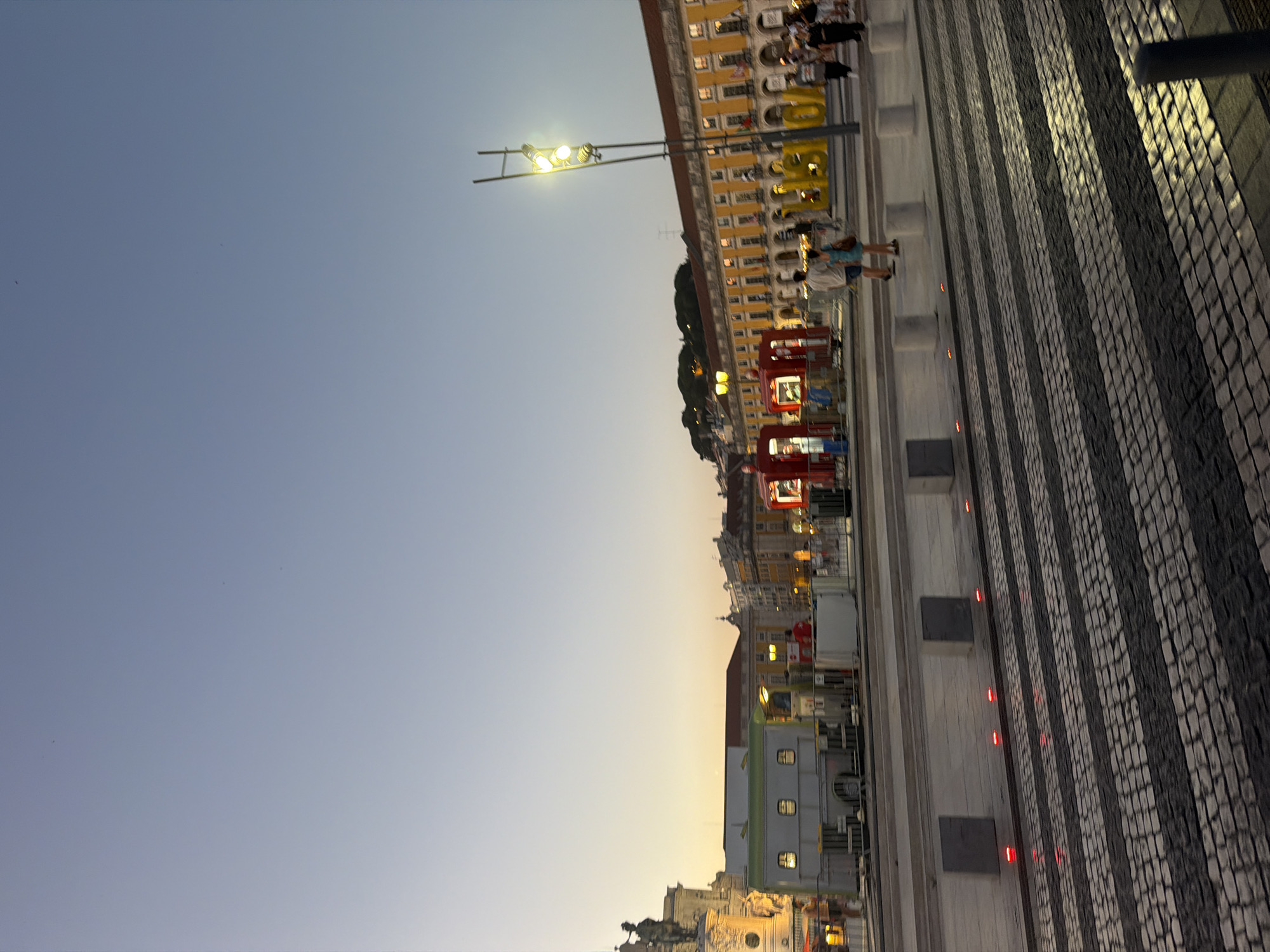 The image depicts Praça do Comércio in Lisbon, Portugal, at dusk. The sky is a gradient of pale blue and warm orange, indicating the setting sun. The square is paved with cobblestones, and a pedestrian crossing is prominently featured in the foreground. Red lights are embedded along the edge of the crossing. Several people are visible, some walking and others standing. To the left, a statue on a pedestal is illuminated. In the middle ground, there are several white portable structures and red temporary booths. A tall lamppost with multiple lights stands on the right side of the image, illuminating the scene. Behind the lamppost, a long yellow building with arched windows and balconies lines the square. A large yellow sign that appears to spell "LISBOA" is visible in front of the building. The overall atmosphere suggests a public gathering or event taking place in the square.