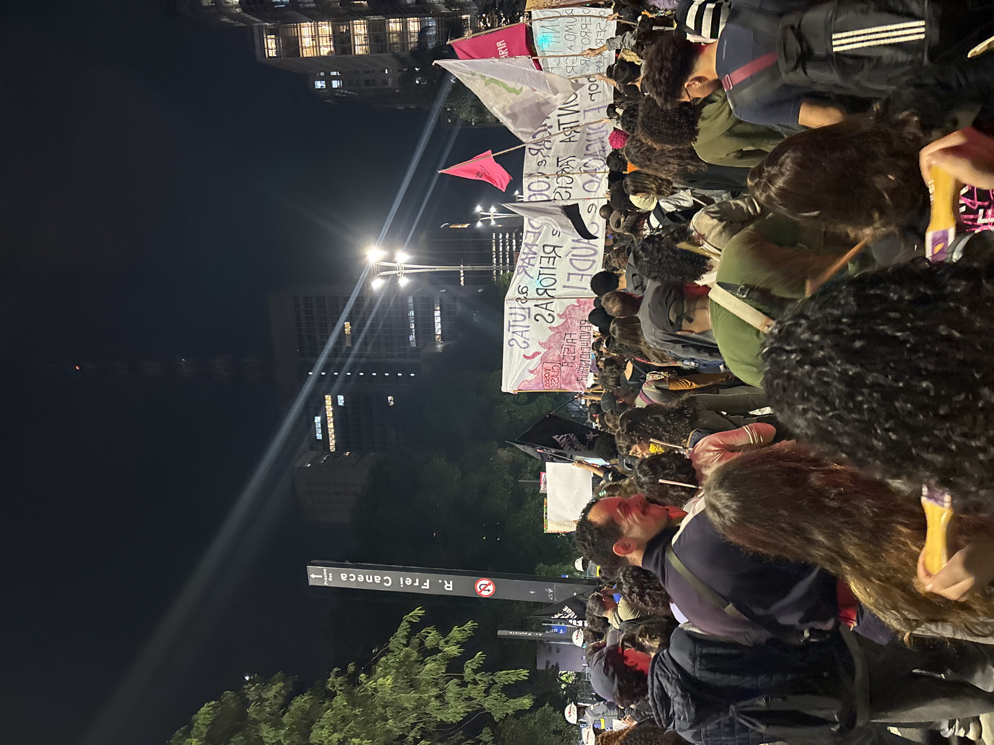The image shows a night scene in São Paulo, Brazil, with a large crowd of people gathered, likely a protest or demonstration. Many are holding signs and banners with messages in Portuguese. In the foreground, a street sign reads "R. Frei Caneca," indicating the location. Buildings and streetlights are visible in the background, illuminating the scene. The crowd is dense, and the atmosphere appears energetic and purposeful.