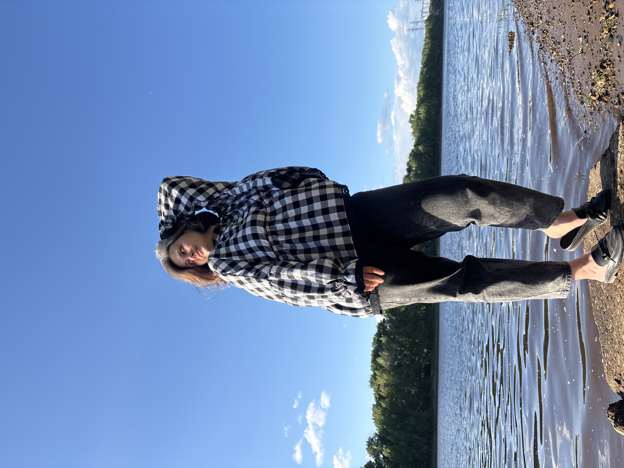 A full-body, low-angle shot shows a woman standing on a rocky shore with her left hand behind her head and her right arm down. She is wearing a black and white checkered blouse, dark gray jeans, and black loafers. The woman is looking to her left, up towards the bright blue sky, with a slight smile. Her brown hair is loose and slightly windswept. Behind her, a large body of water stretches out, with a tree-lined shore in the distance. The sky is mostly clear with a few scattered white clouds. The overall scene depicts a serene, outdoor setting, likely near a river or lake, on a sunny day.
